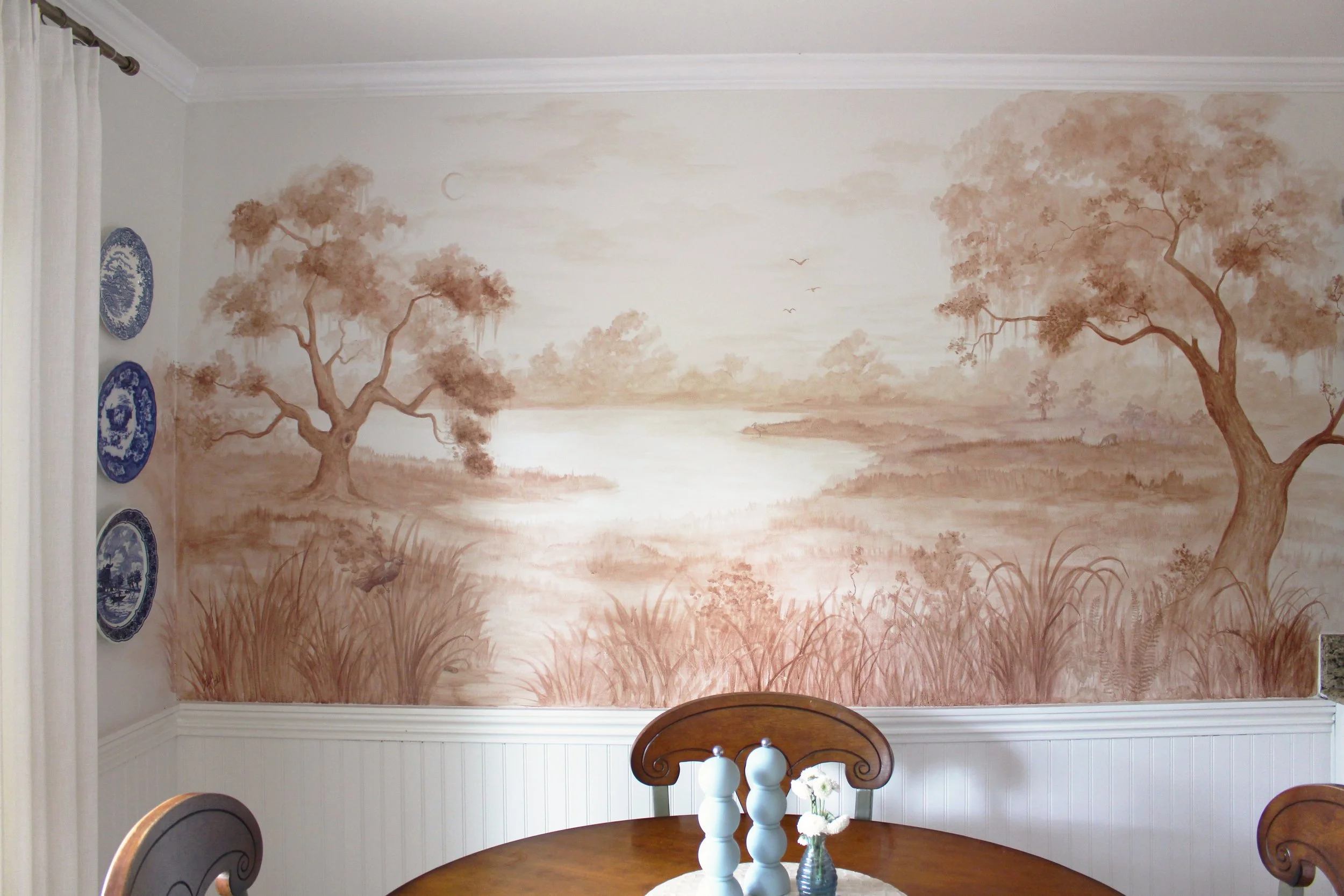 A dining room wall mural depicting a peaceful nature scene with trees, a river, birds, and a crescent moon painted in shades of brown and beige.