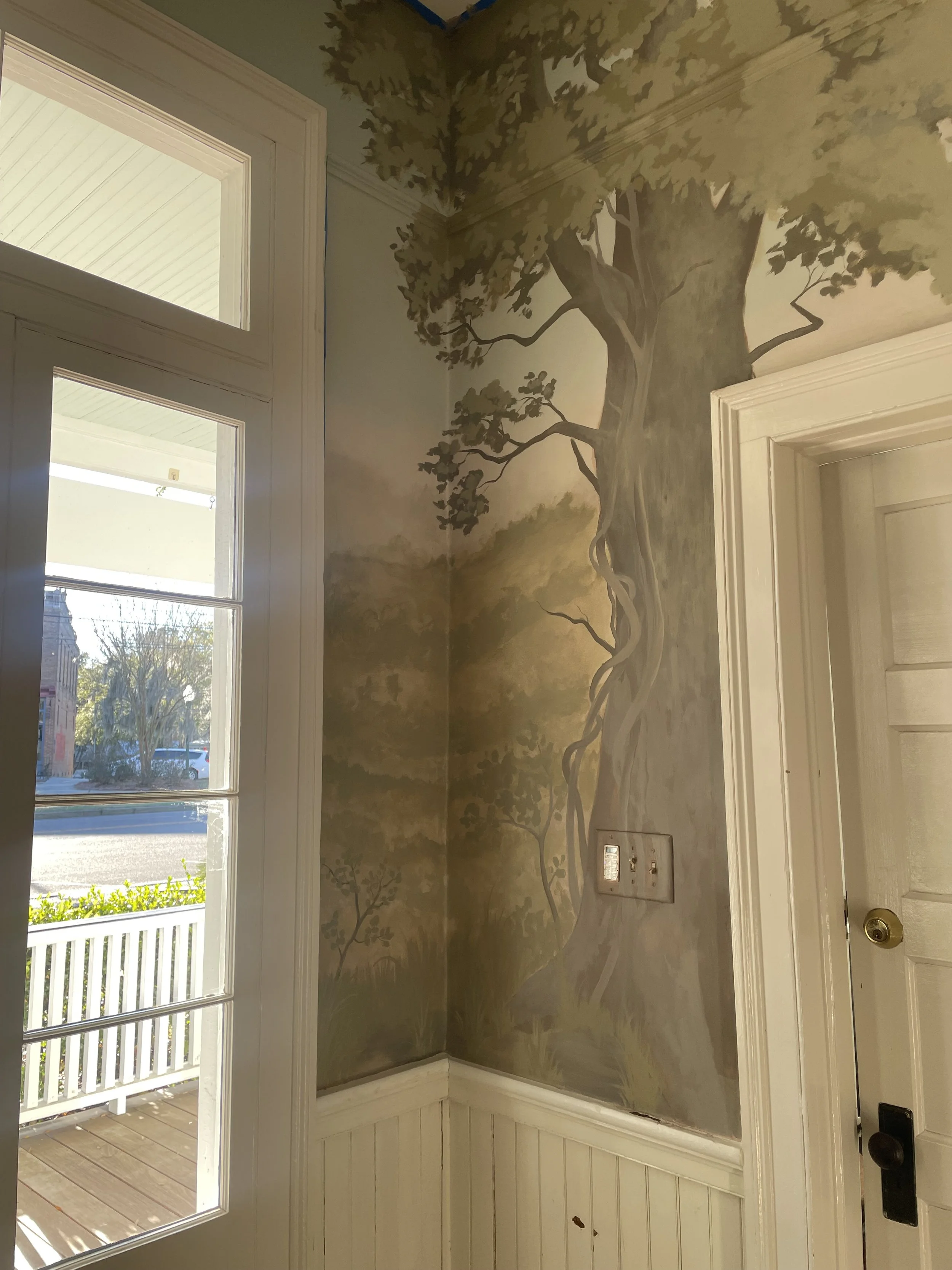 Interior corner of a room with a painted mural of a tree on the wall next to a window and a door.