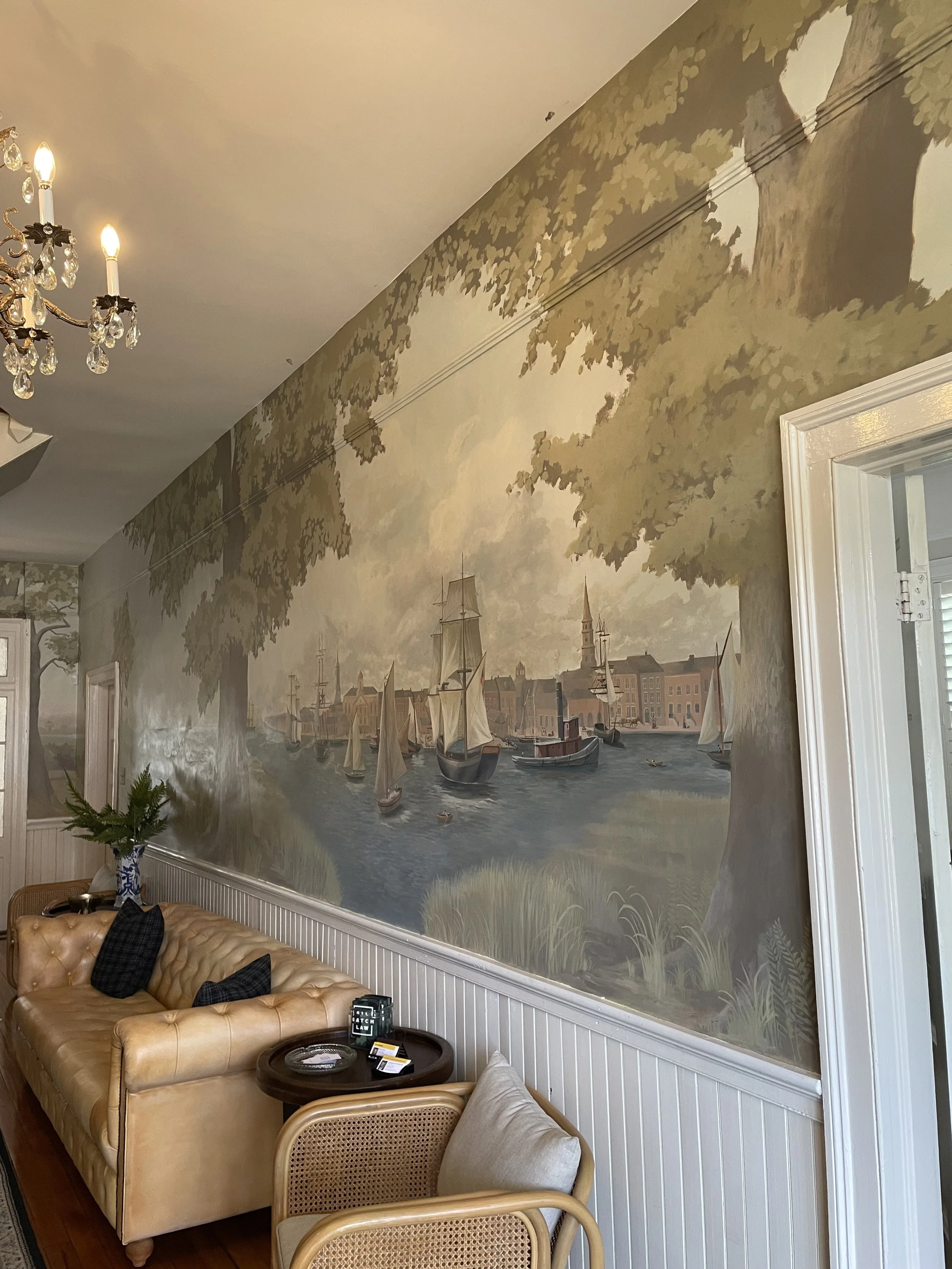 Interior of a room with a large mural of historic Charleston harbor with sailboats and buildings, a tan leather sofa, a small round table with items on it, a wicker chair, and a chandelier with crystal adornments.