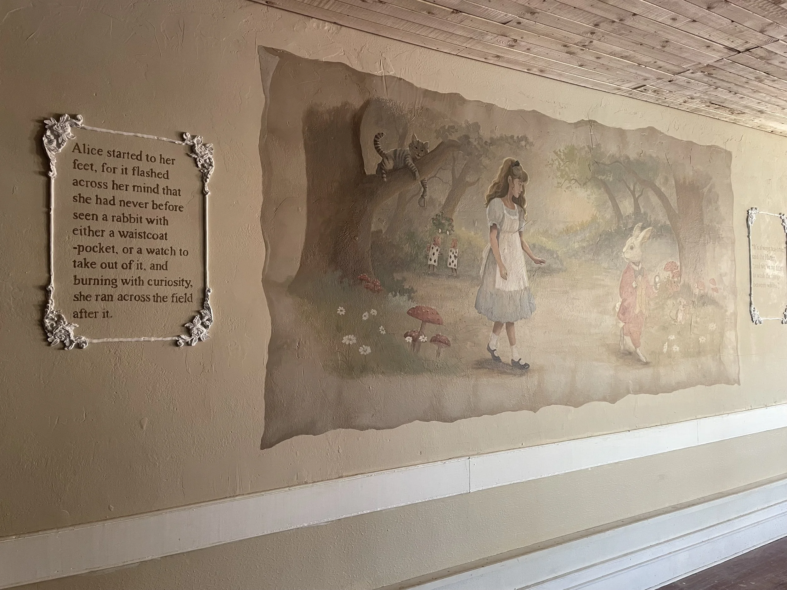 Custom mural of Alice in Wonderland in a garden with a cat on a tree branch, a rabbit, and other whimsical characters. There is also a decorative rococo relief frame with a quote about Alice.