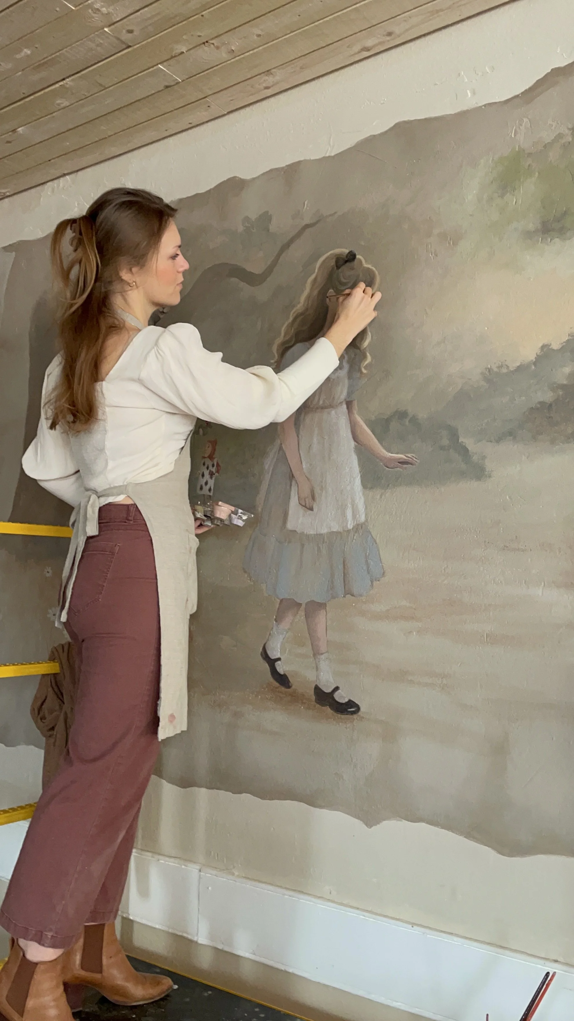 Charleston mural artist painting a mural of a girl with long blonde hair, a dress, and black shoes on a wall.