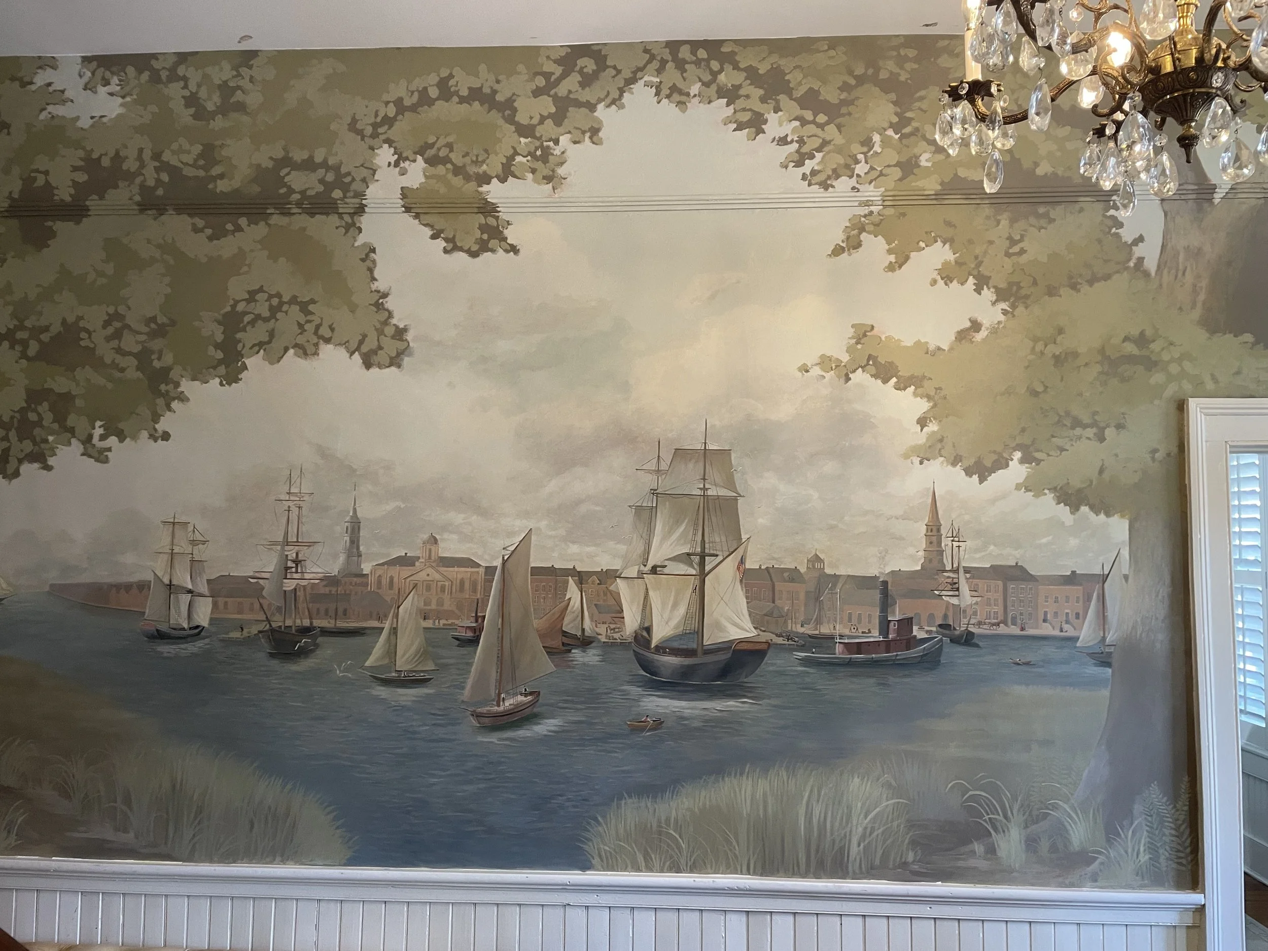 A wall mural illustrating a historic Charleston harbor scene with sailing ships and buildings, topped with a decorative chandelier and ceiling paint pattern.