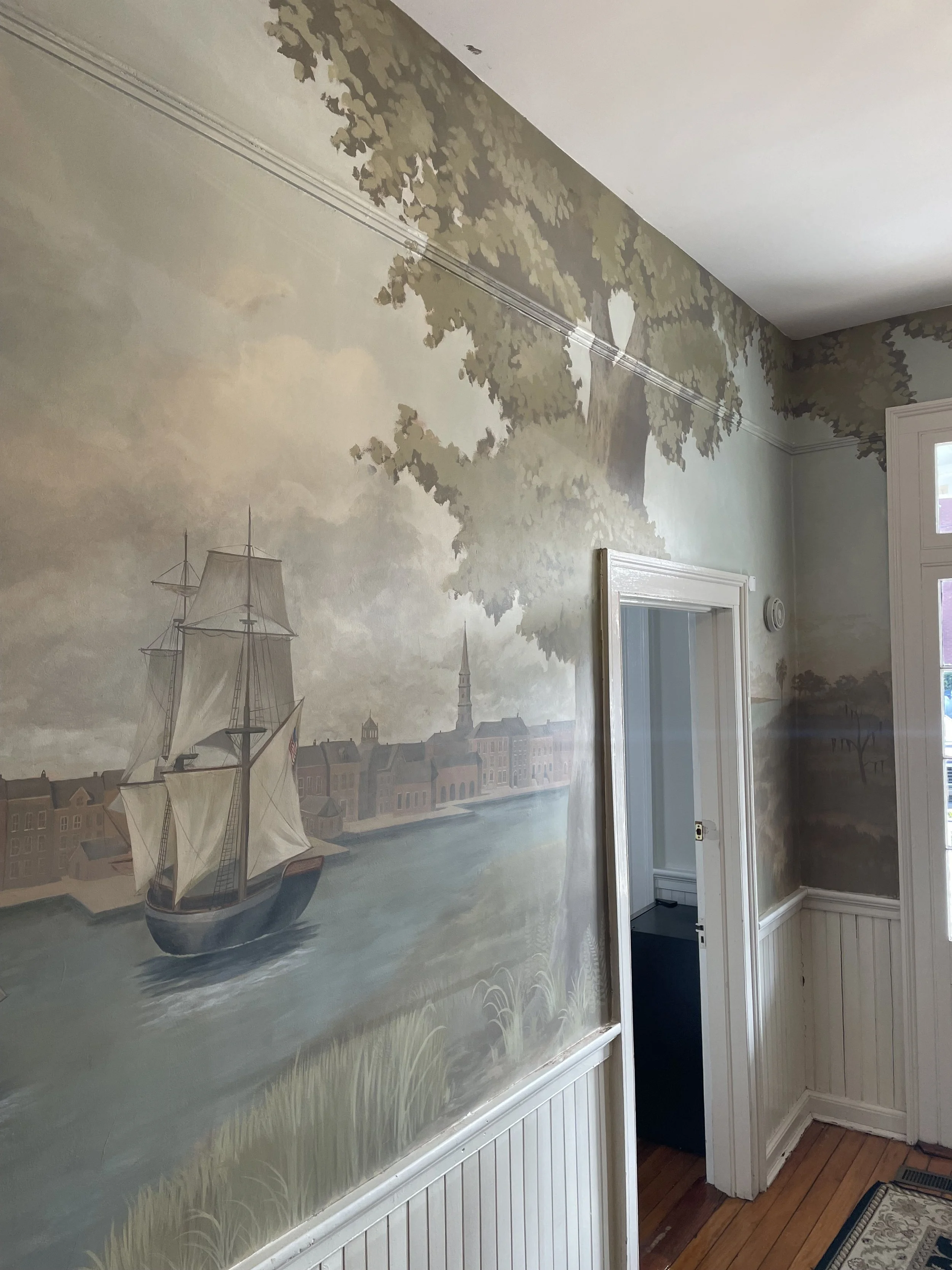 Interior wall mural depicting a river scene with a sailing ship, historic buildings, and a church steeple, framed by green trees and foliage, in a house with wooden flooring.
