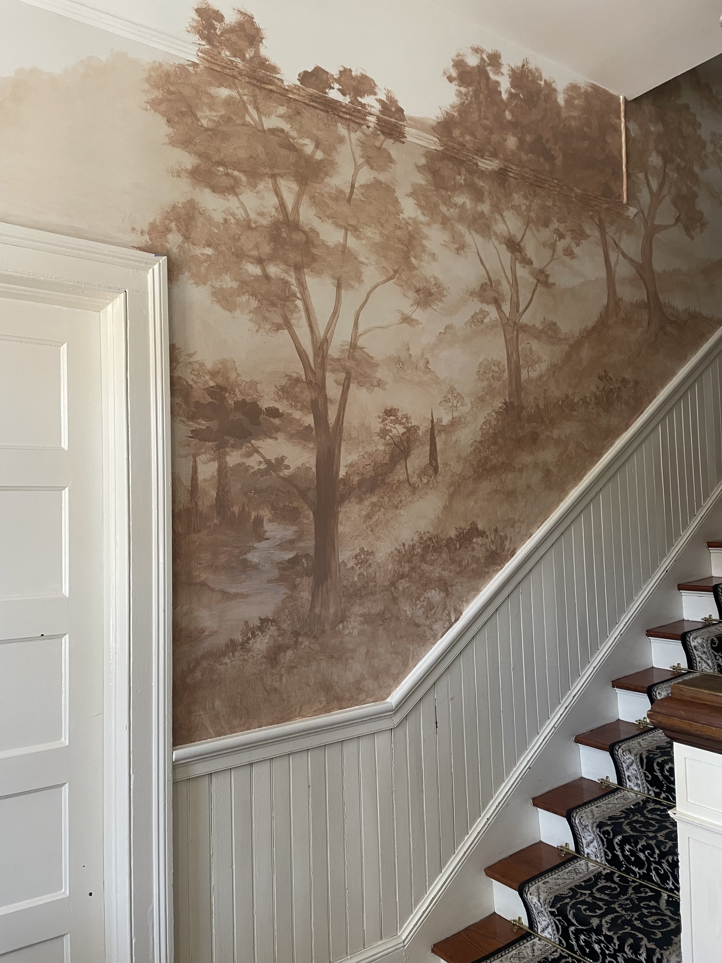 Faux landscape mural of trees and a river on the wall beside a staircase inside a house by professional artist Gabrielle Hasenstab.
