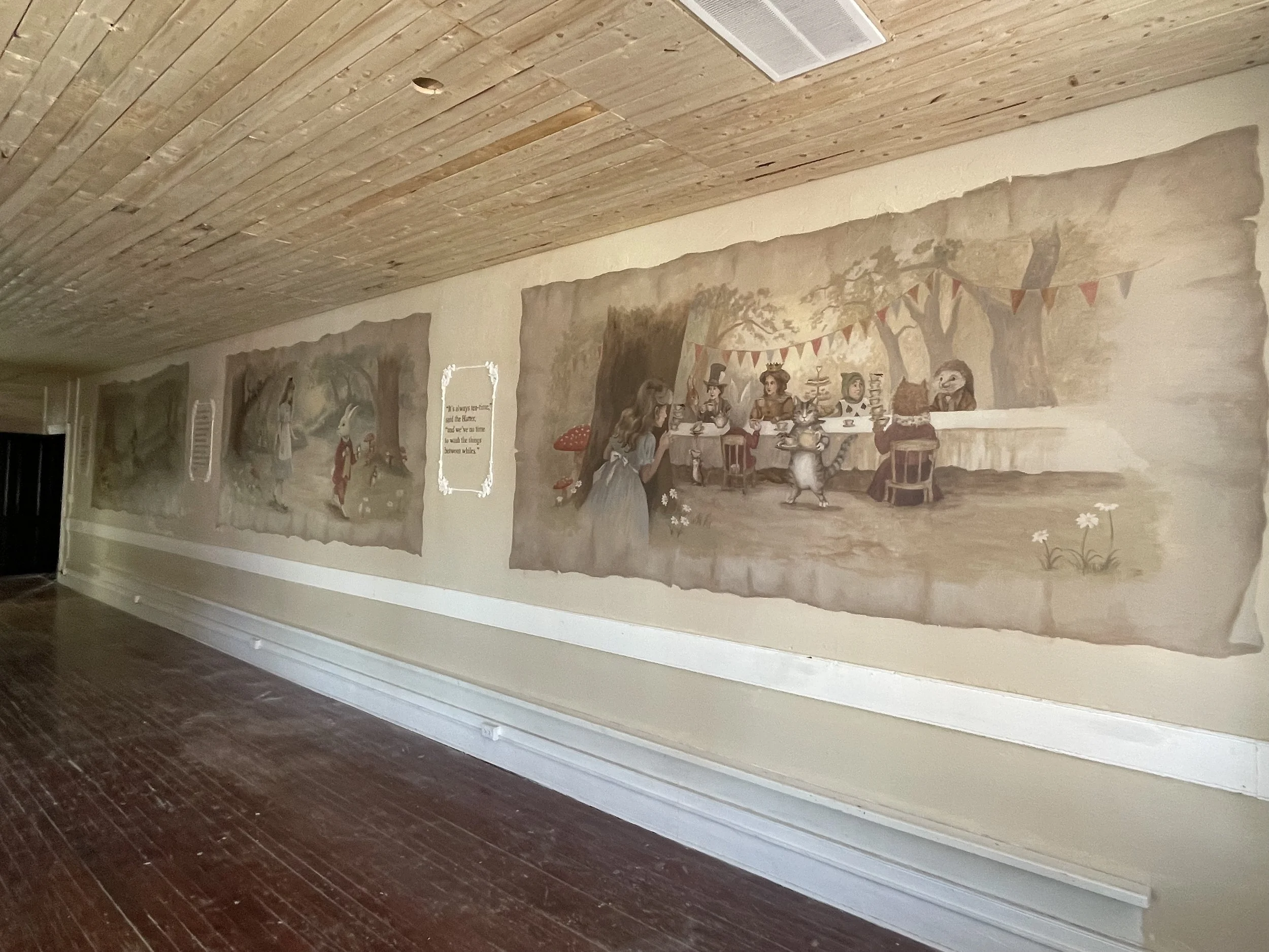 A painted mural on a beige wall depicting Alice in Wonderland characters, including Alice, the Mad Hatter, the White Rabbit, and the Cheshire Cat, with trees and a garden scene in the background.
