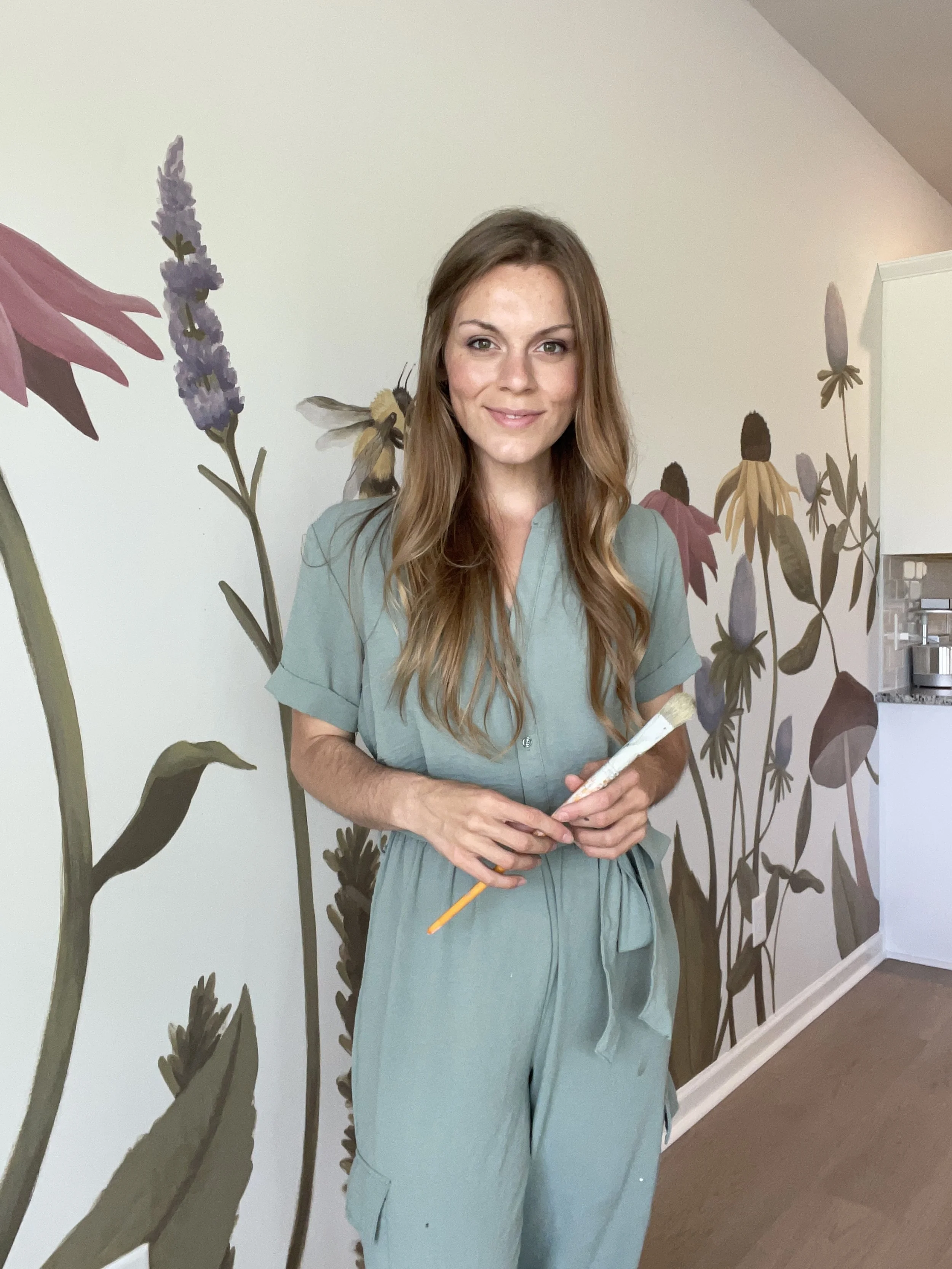 Charleston mural artist Gabrielle Hasenstab stands in front of a mural she painted of wildflowers wearing a light green jumpsuit, smiling and holding a paintbrush.