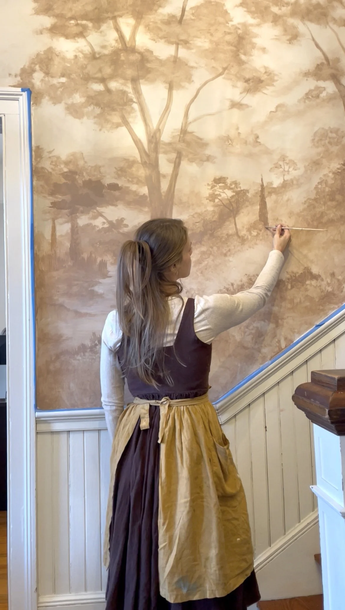 Female professional artist Gabrielle Hasenstab  painting a mural of trees on a wall in a cozy interior space.