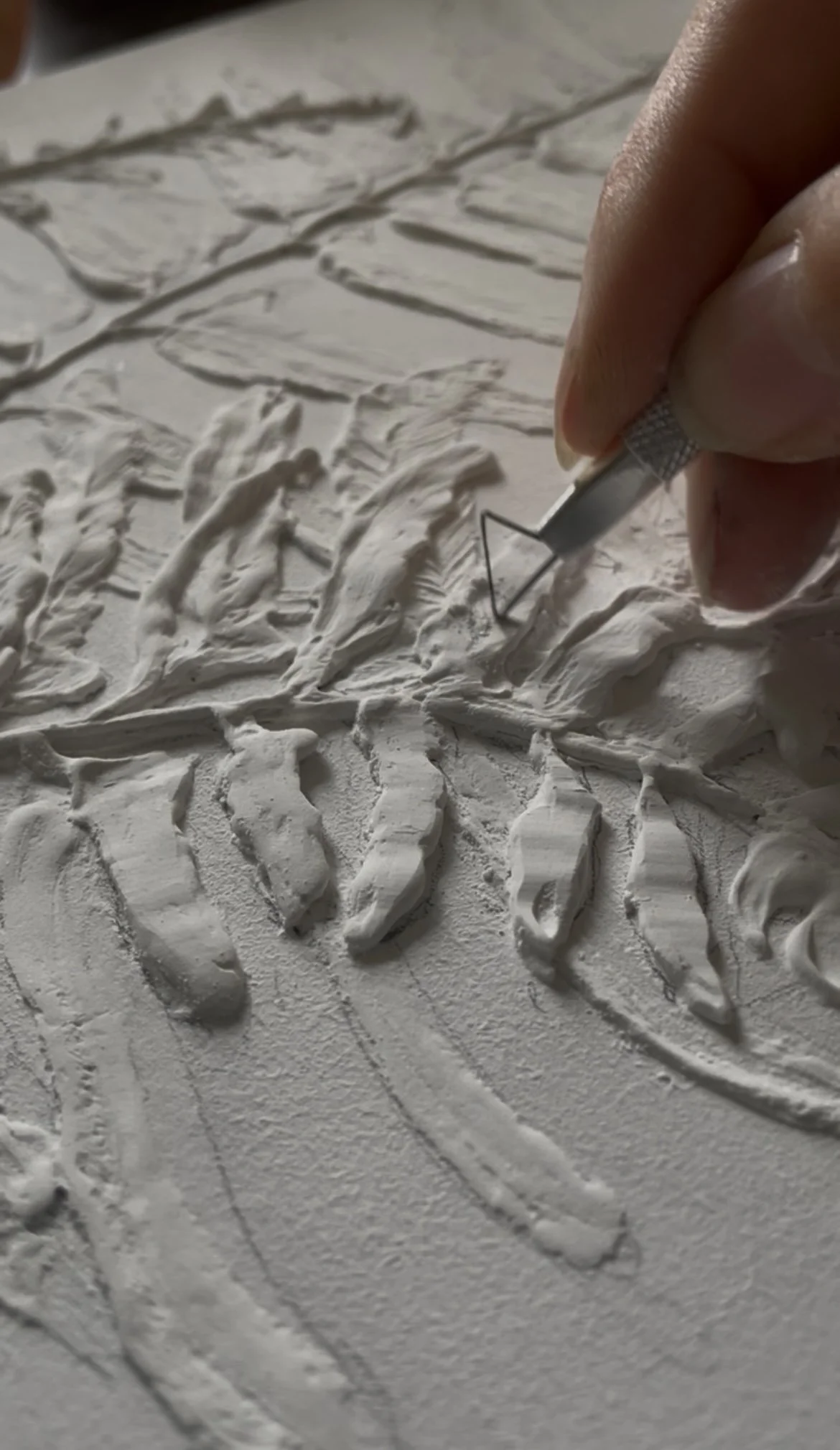 Close-up of an artist carving a detailed piece of white clay or plaster with a metal sculpting tool, creating intricate textures and patterns.
