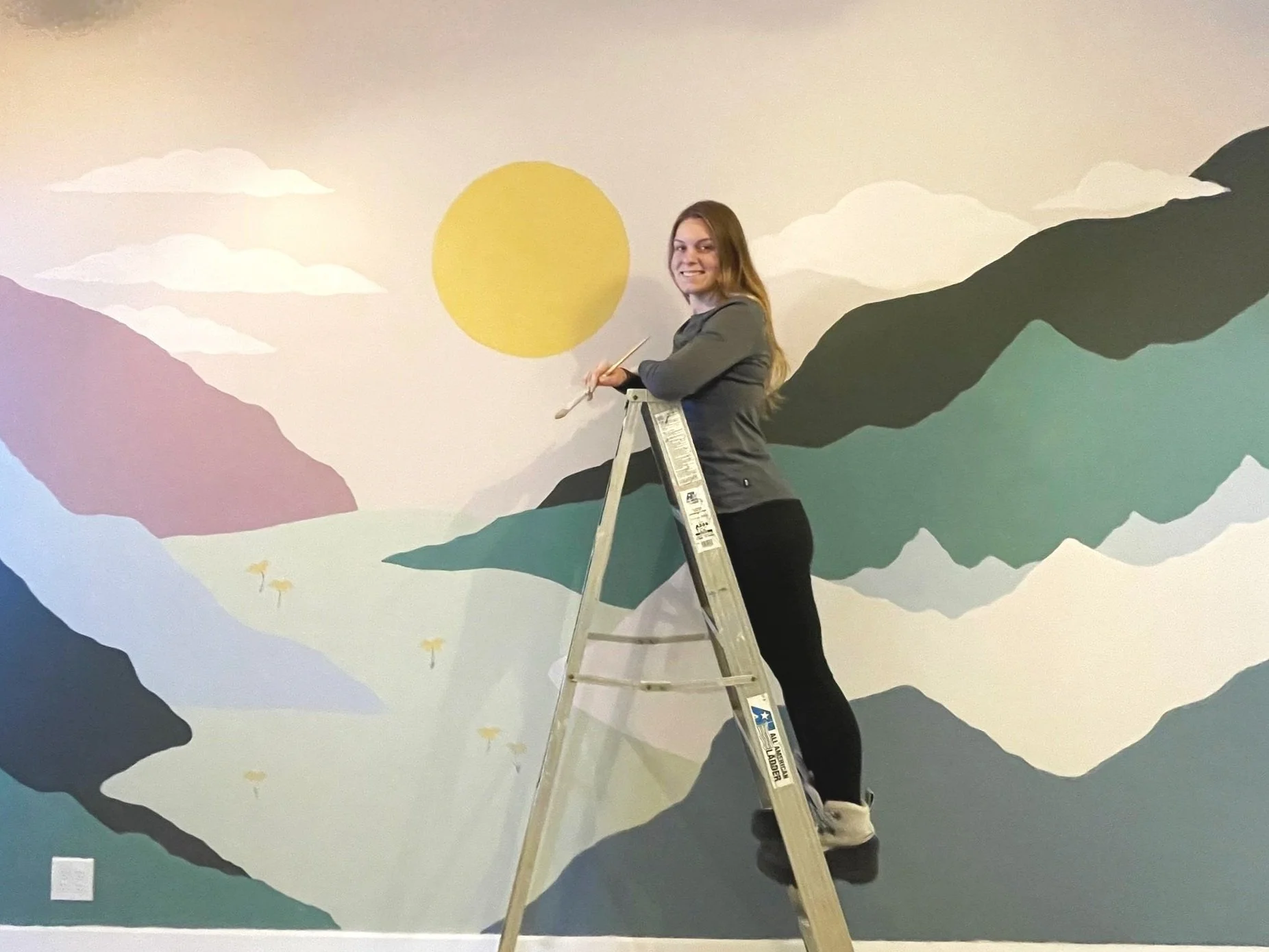 Mural artist Gabrielle Hasenstab standing on a ladder painting a mural of mountains, a yellow sun, and clouds on a wall.