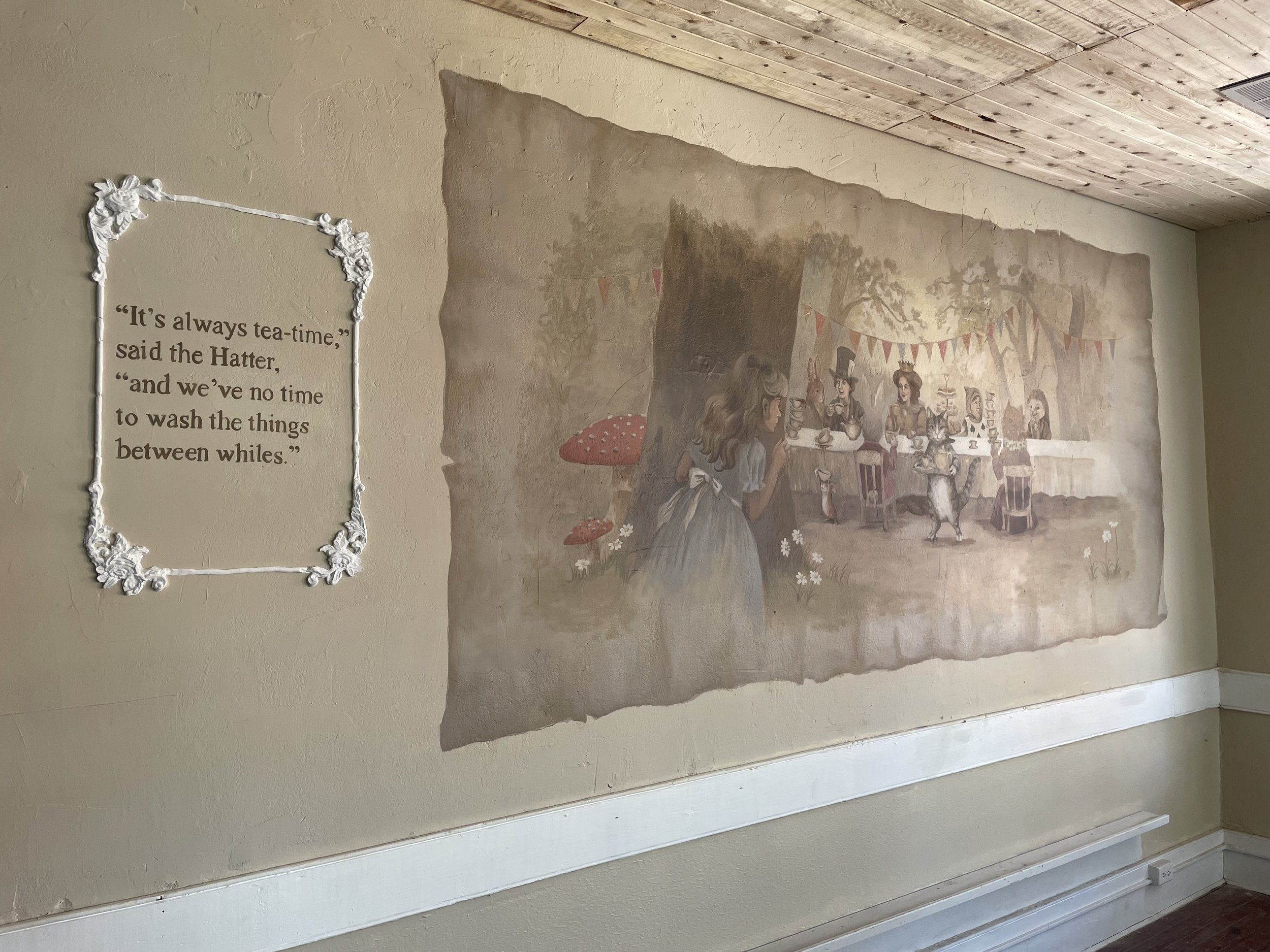 Wall mural depiction of a whimsical outdoor gathering with characters, animals, and colorful banners, with a quote about tea in a decorative frame.