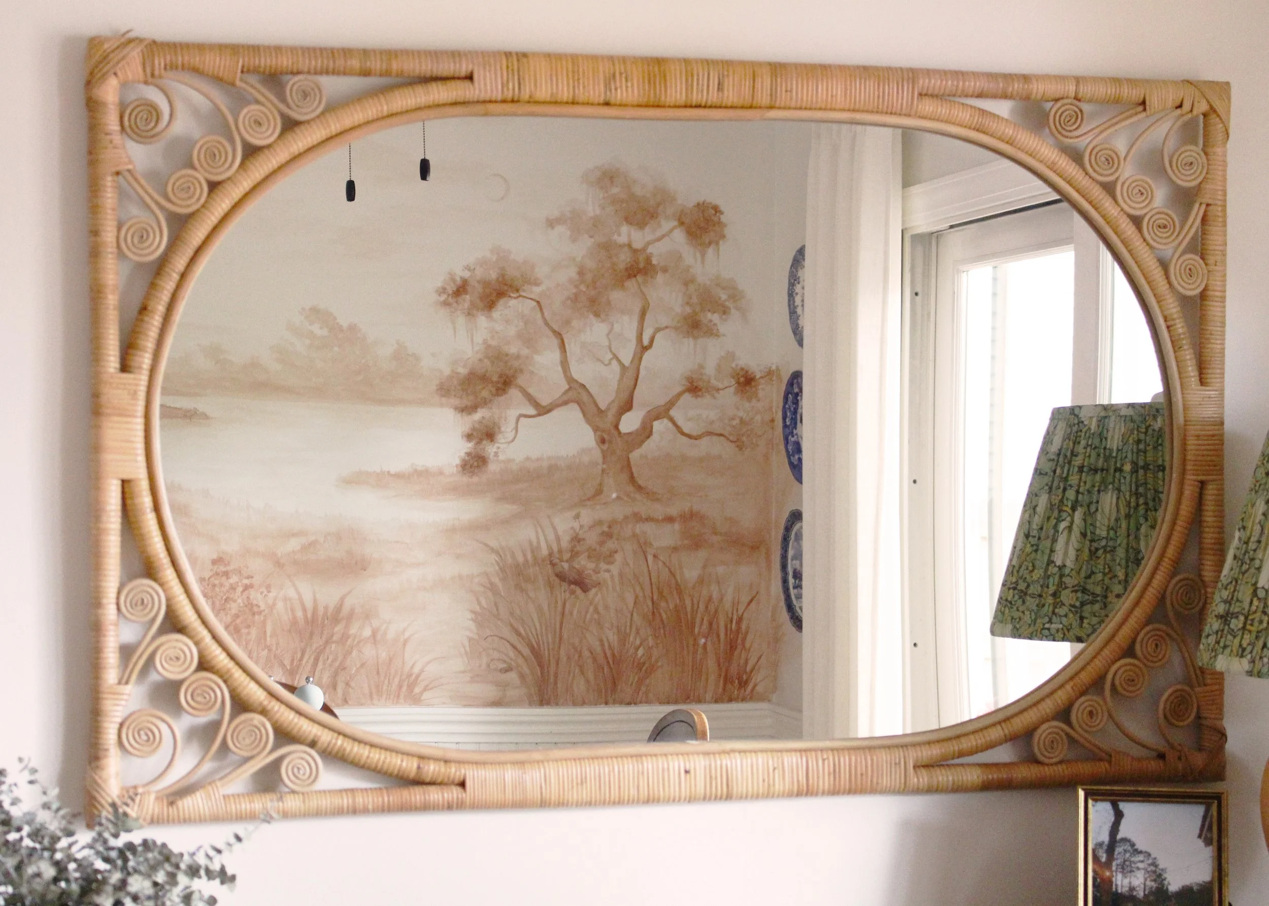 Decorative mirror with a woven rattan frame reflects a living room with a wall mural of a tree, a window, a patterned chair, and a framed picture.