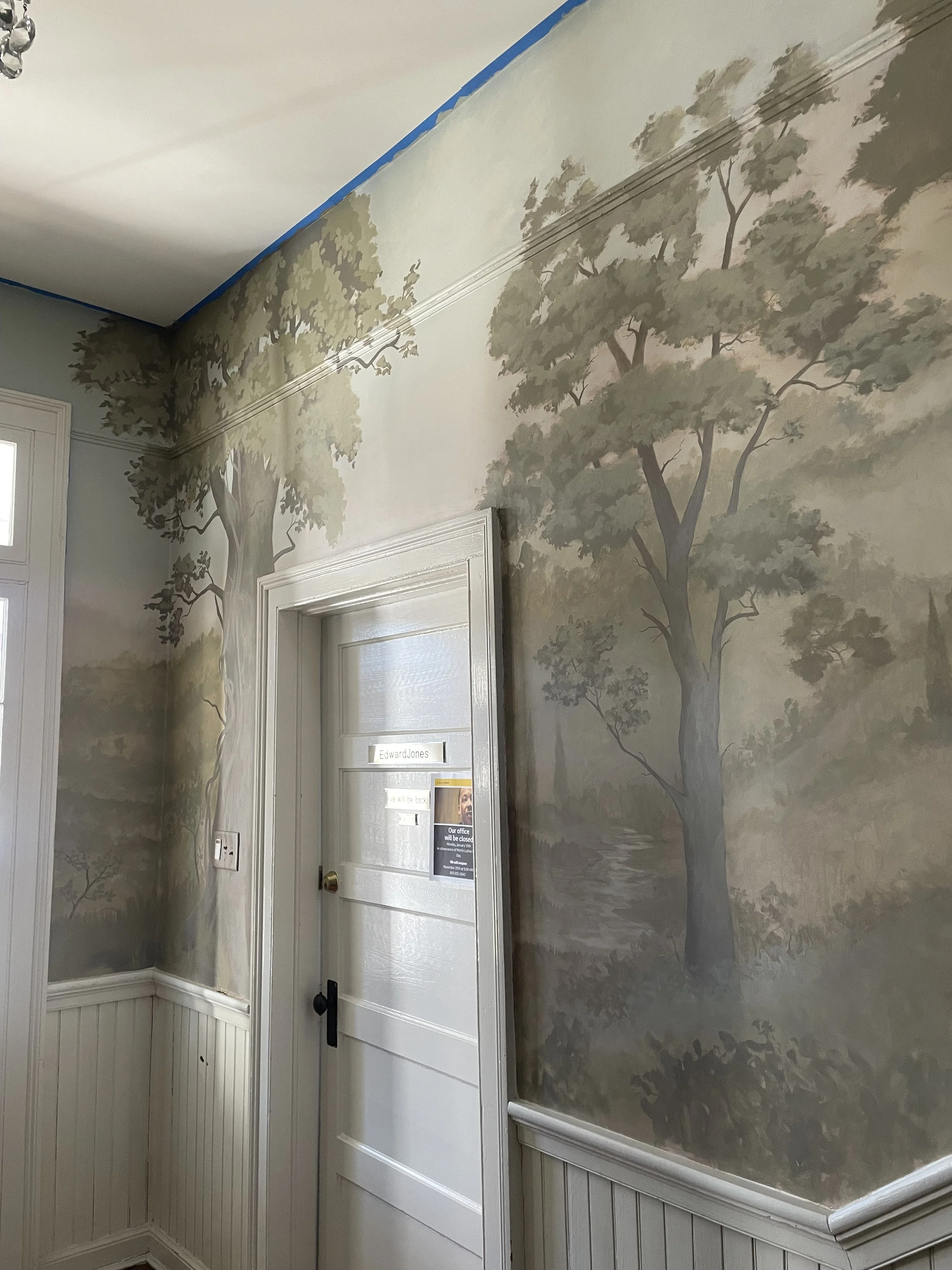 Interior wall with a painted scenic mural of trees and a landscape in muted earthy tones, featuring a white door with signs and a window to the left. The ceiling is partially covered with blue painter's tape.