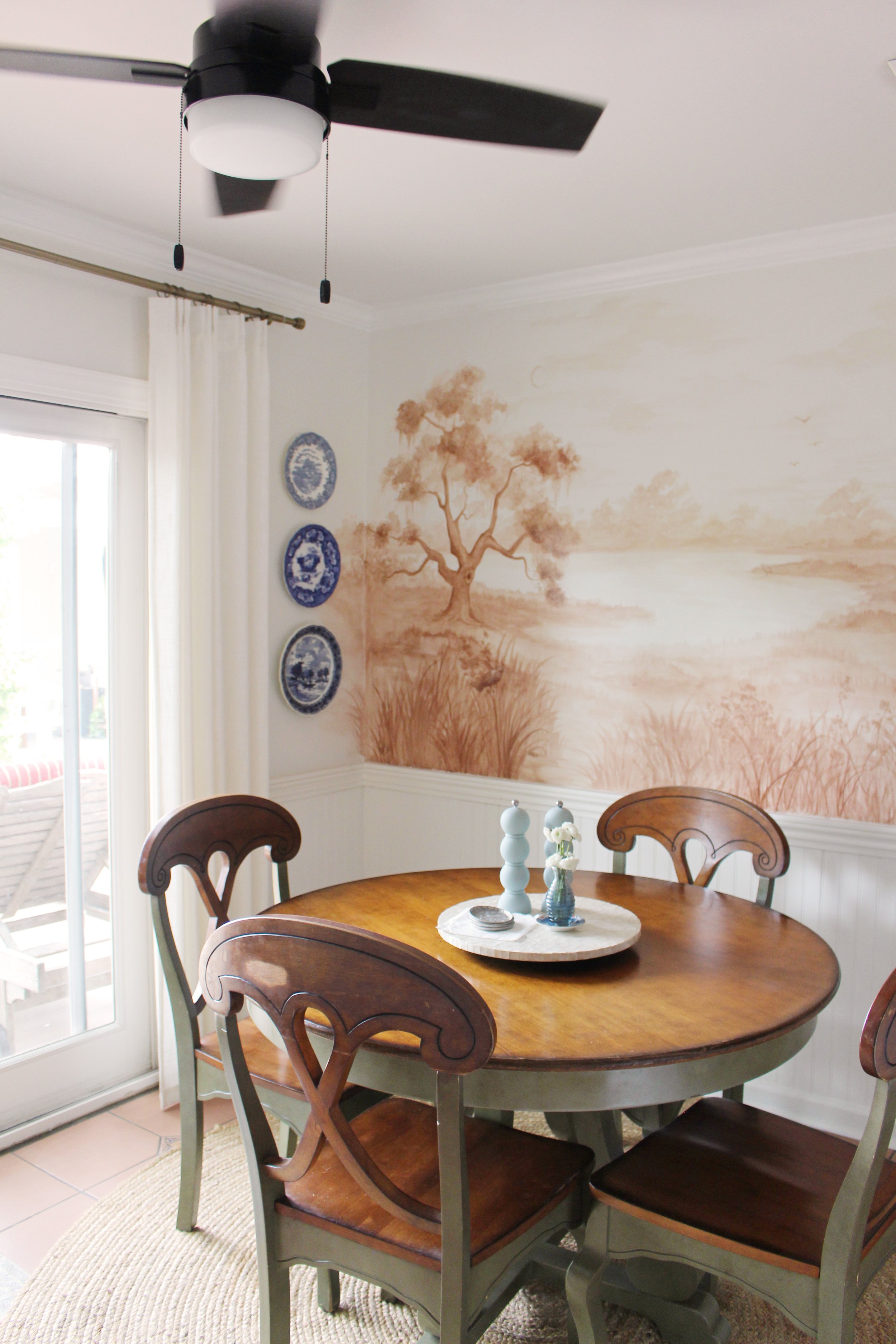 A dining room with a wooden table surrounded by matching chairs, a ceiling fan overhead, a wall mural of a tree and landscape, and a sliding glass door with white curtains.