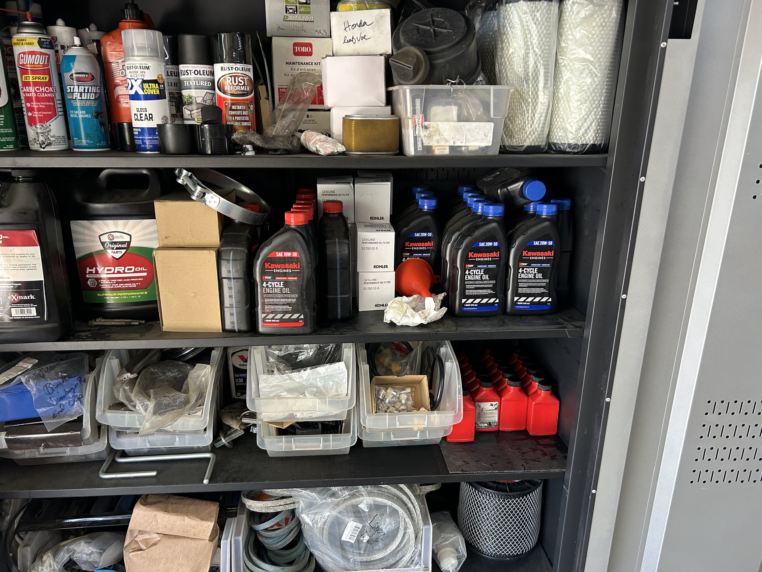 Shelves filled with various automotive and garage supplies, including engine oil bottles, spray cans, plastic containers with small parts, cardboard boxes, and filters.