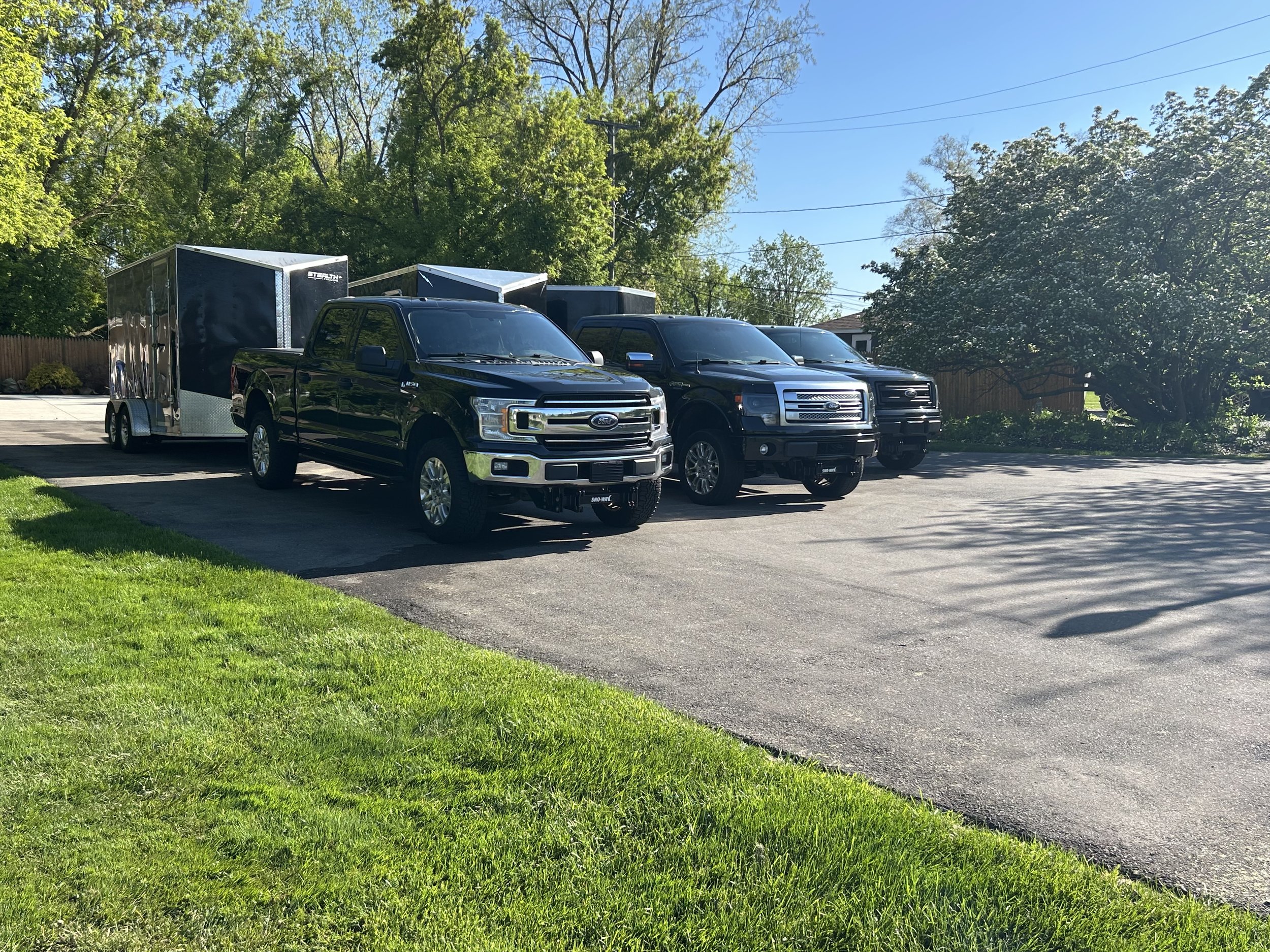 Three black pickup trucks, one towing a black trailer, parked on a paved driveway surrounded by green grass and trees.