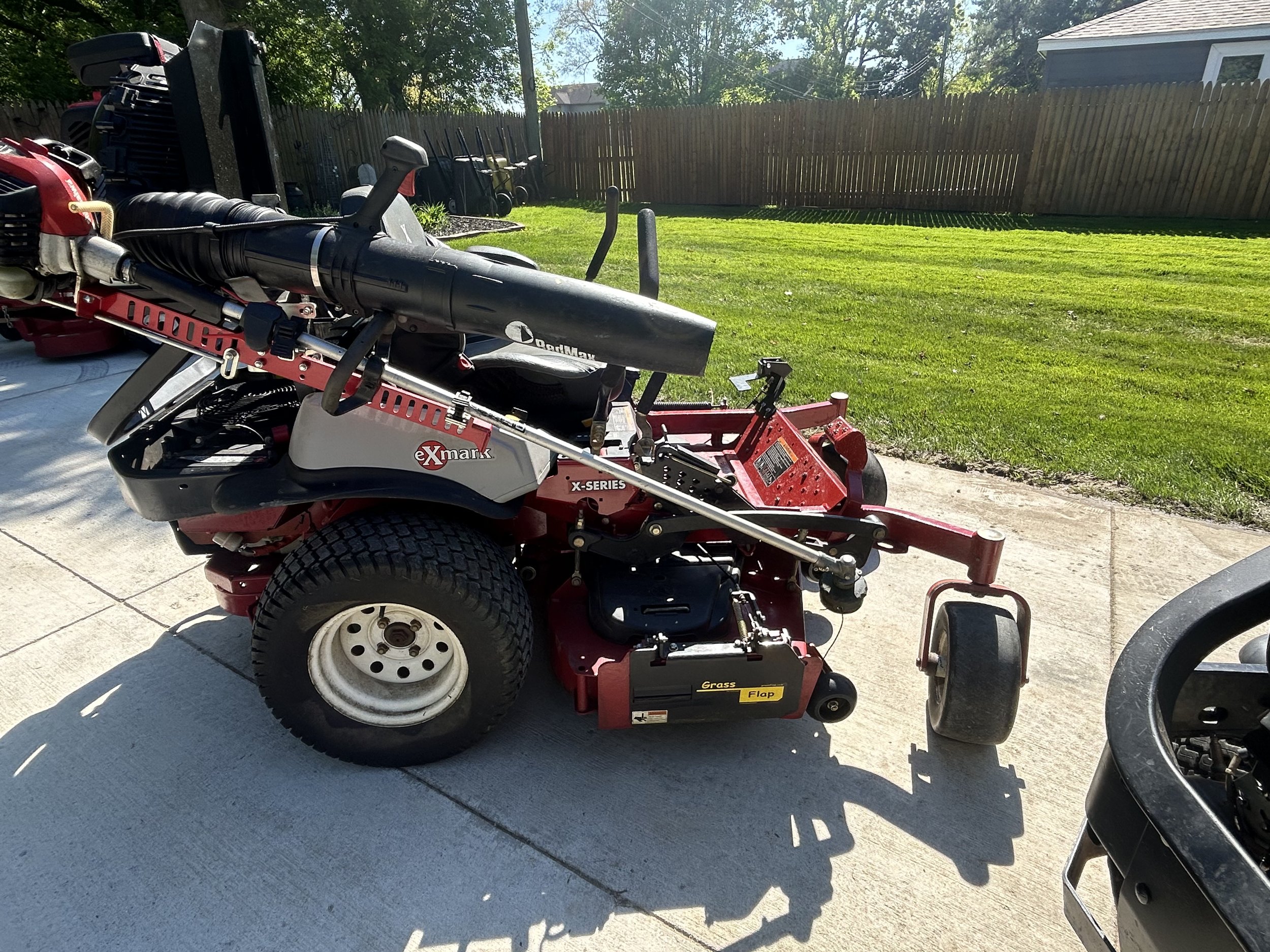 Zero-turn lawn mower parked on a concrete driveway with a green lawn and wooden fence in the background.