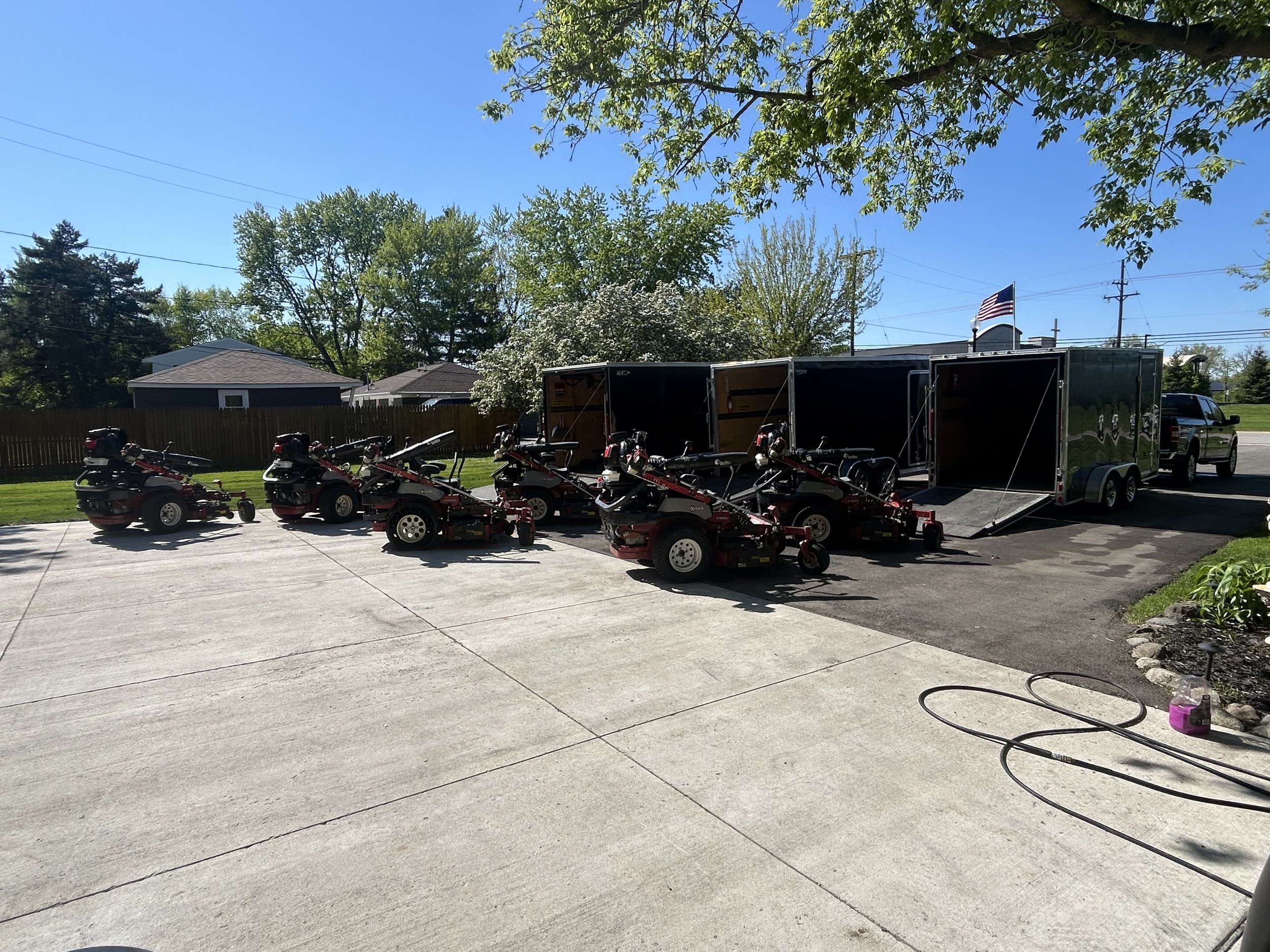 Several red and black lawn mowers parked on a driveway, next to large trailers and a truck with an American flag. Trees and houses in the background.
