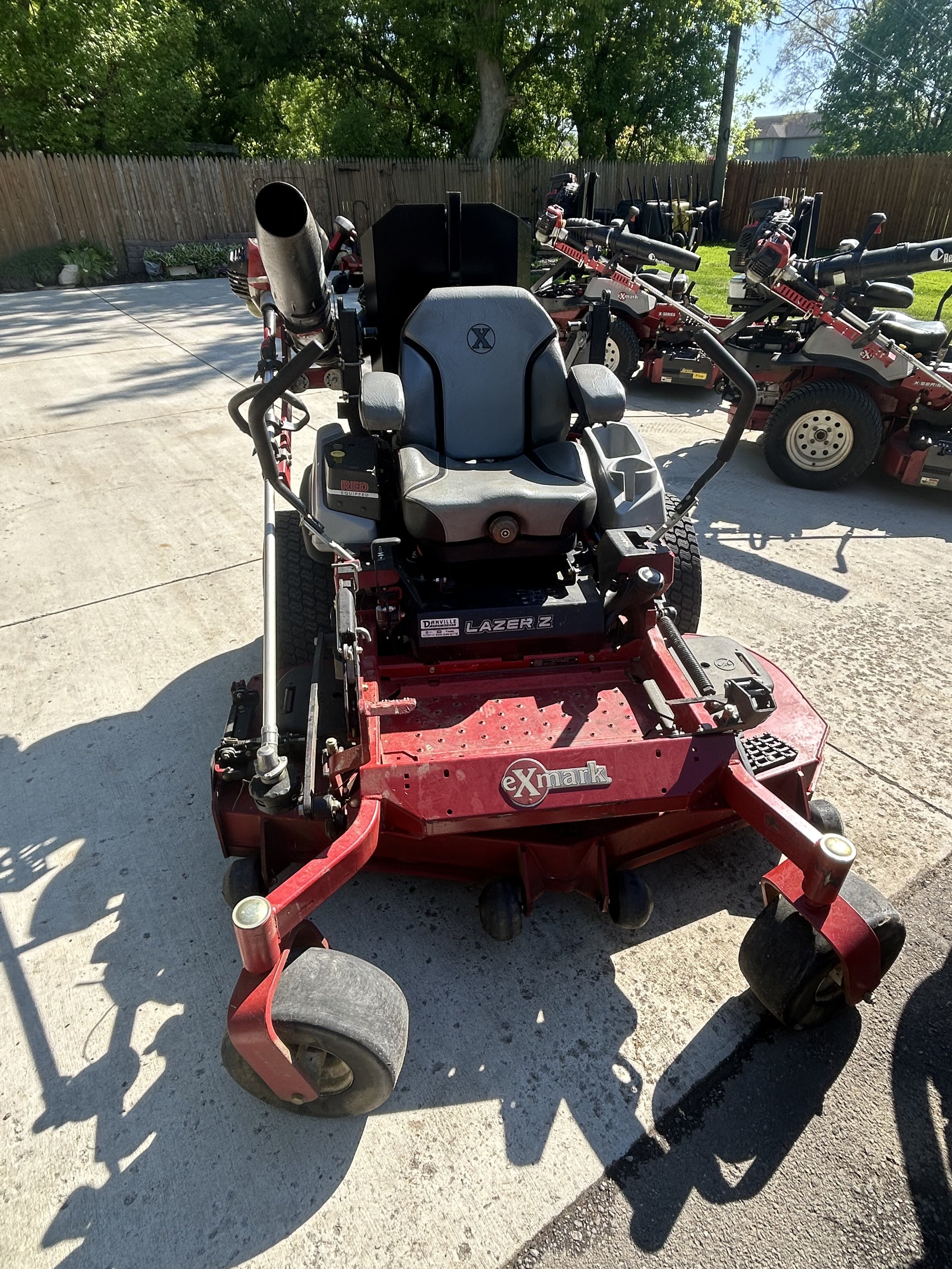 Red Exmark Lazer Z riding lawn mower on pavement, surrounded by other lawn equipment, and a wooden fence in the background.
