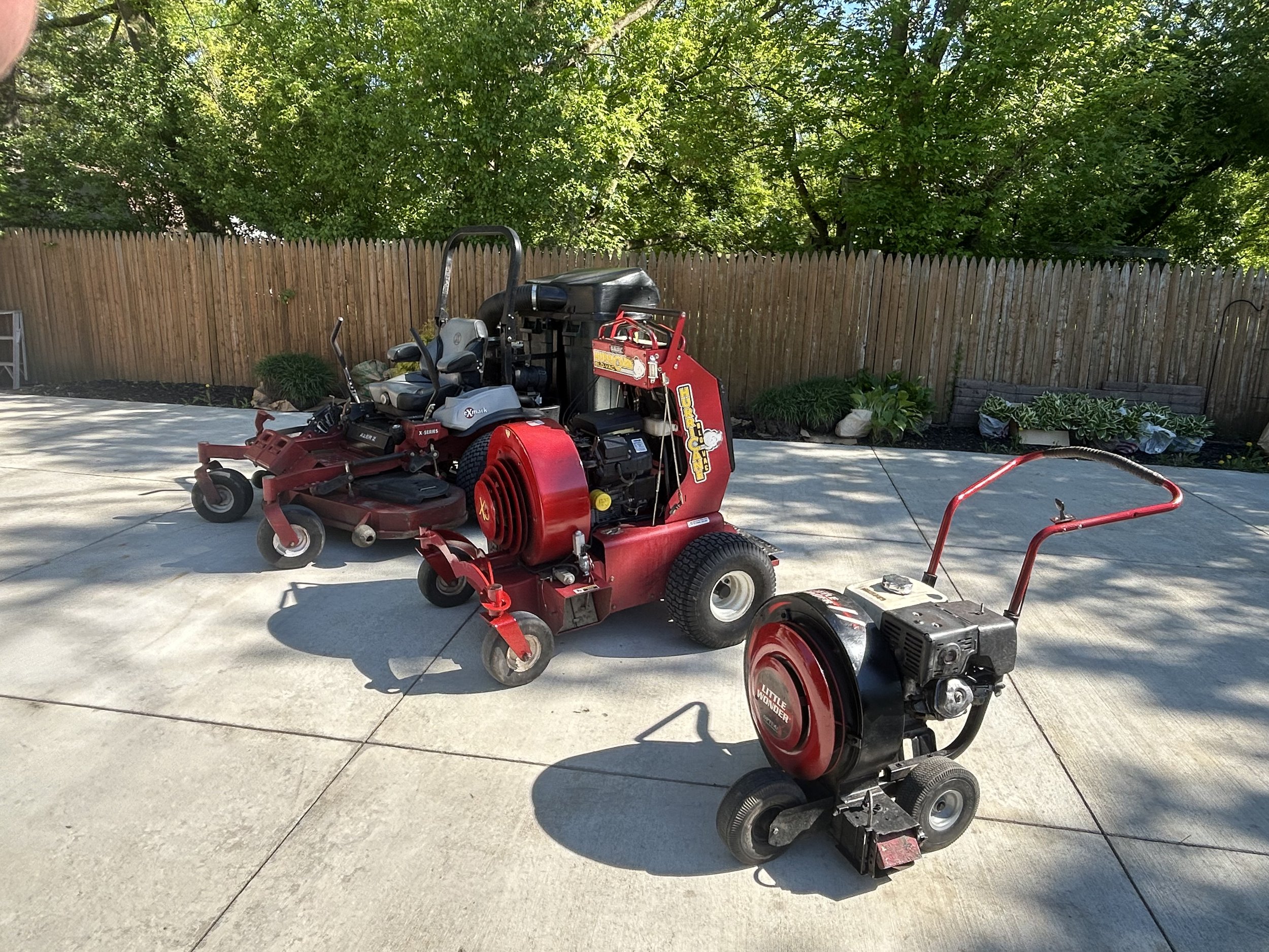 Lawn equipment including a red zero-turn mower and a small leaf blower on a paved surface with trees and a wooden fence in the background.