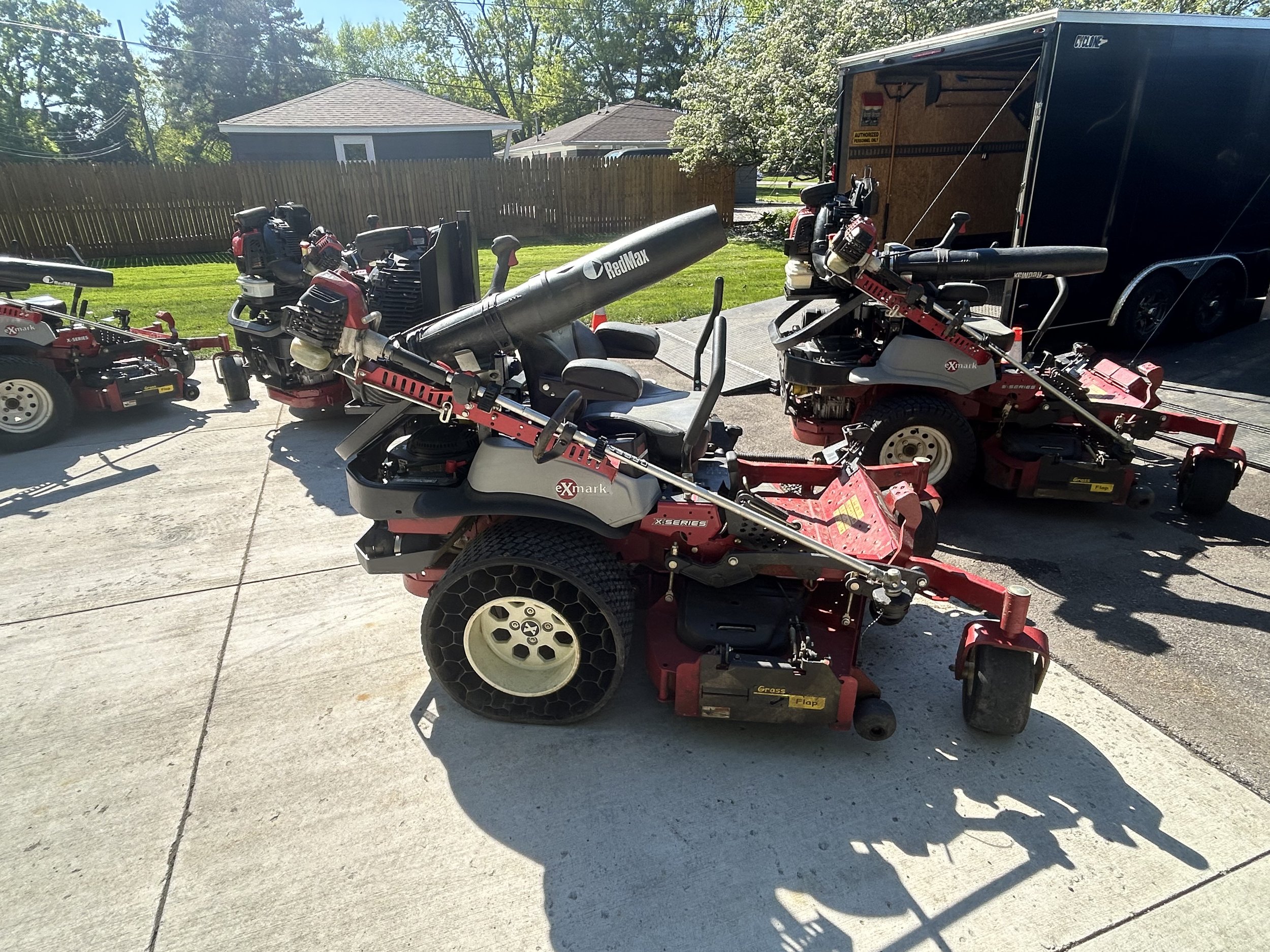 Several red zero-turn lawn mowers parked on a concrete driveway with a trailer in the background.