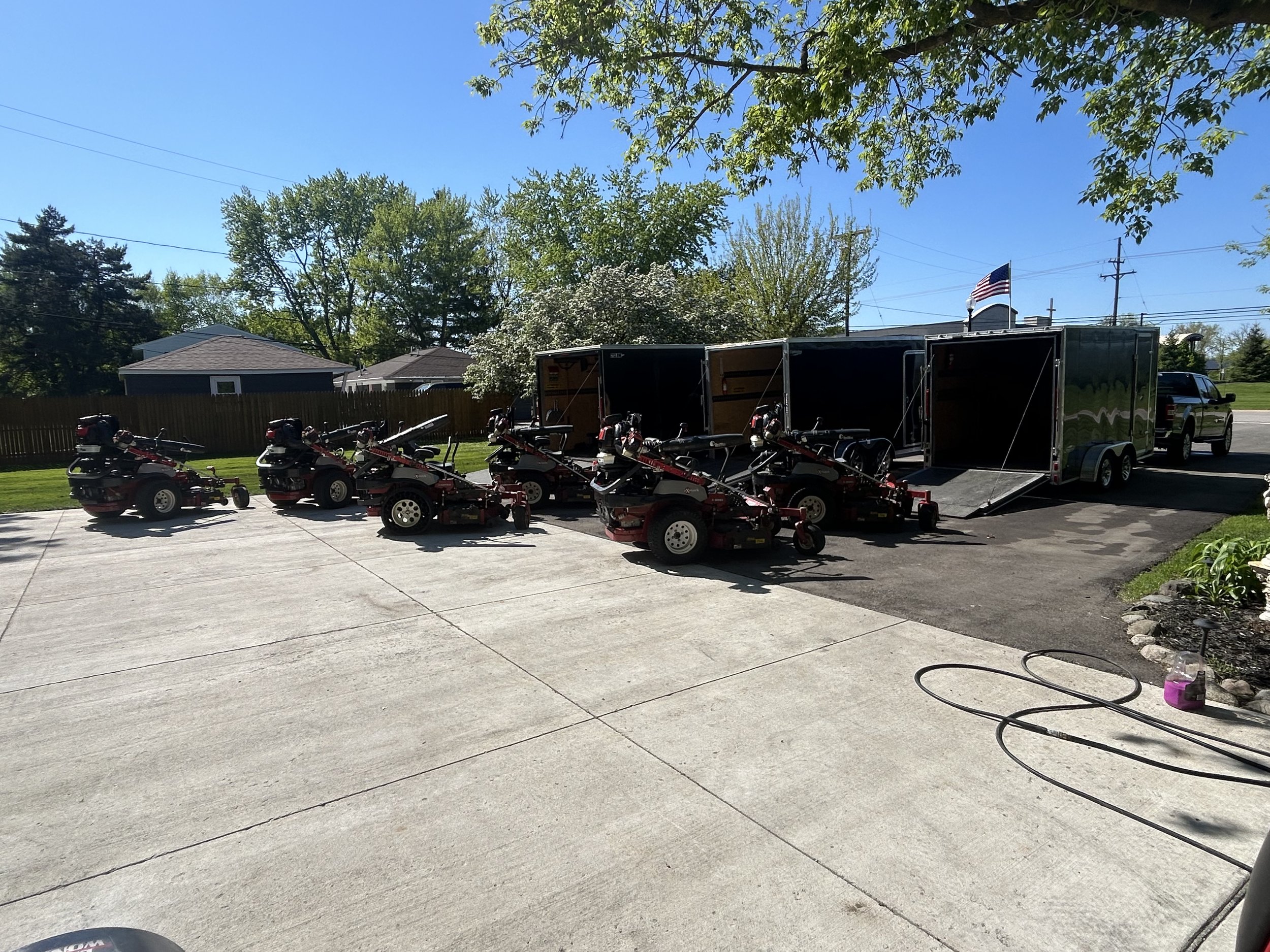 Commercial riding lawn mowers parked on a driveway near trailers and a pickup truck, with trees and a clear blue sky in the background.