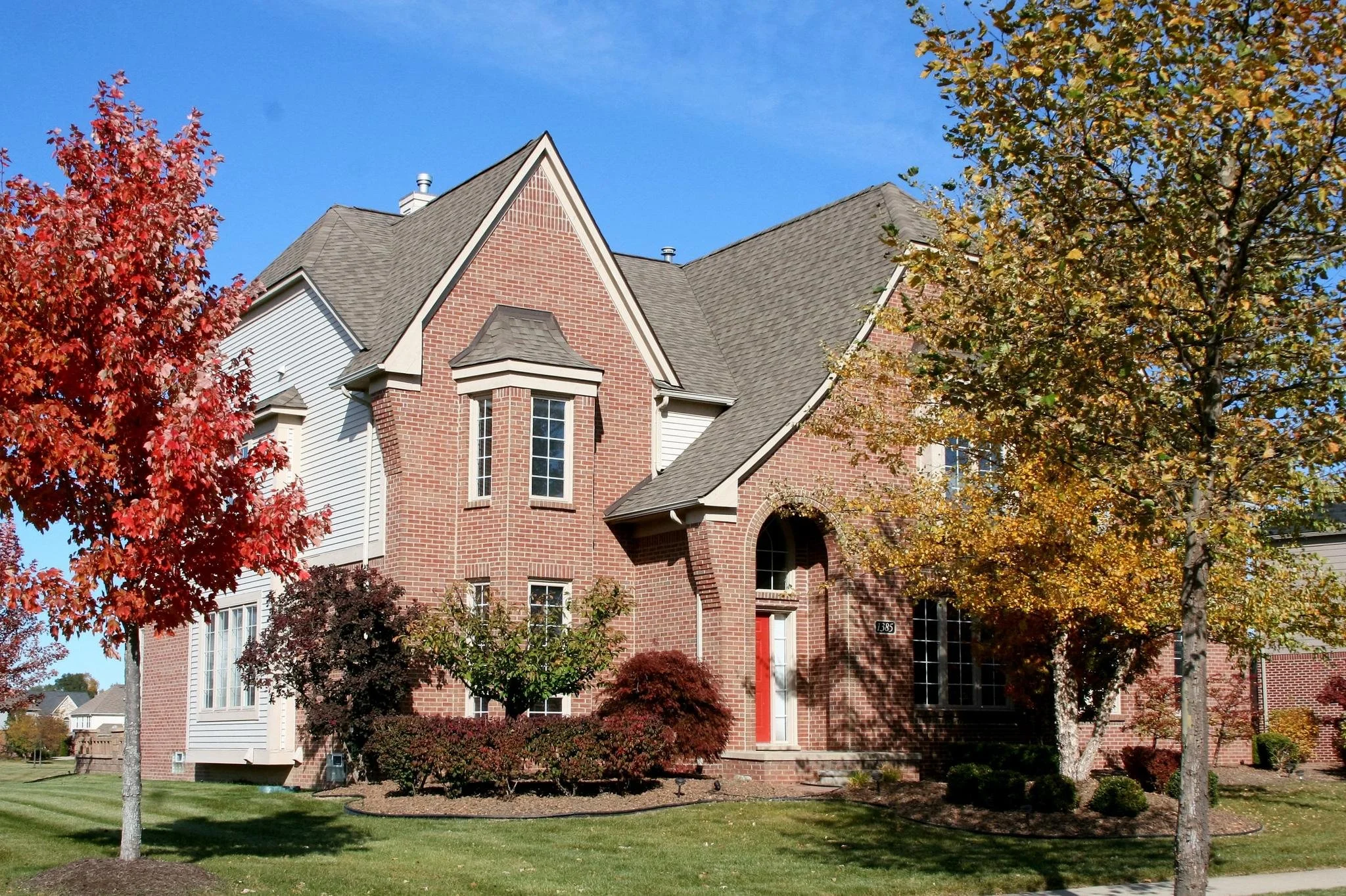 Red brick house with peaked roof, surrounded by fall trees with red and yellow leaves, and a well-maintained lawn.