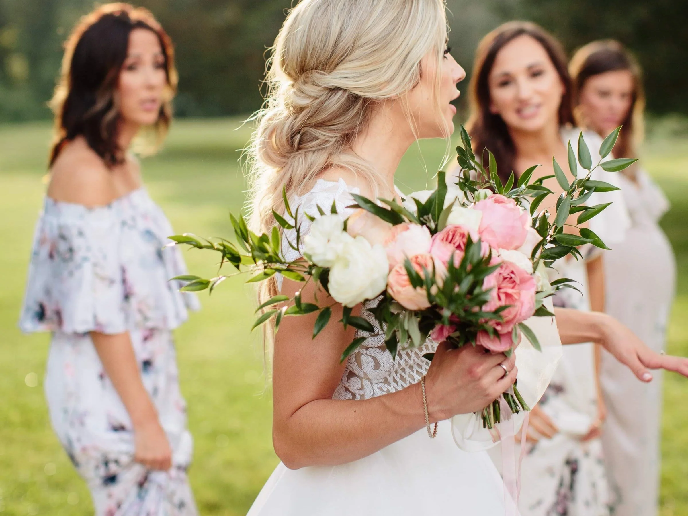 A bride holding a bouquet of pink and white flowers, with three women standing in the background outdoors in a grassy area.