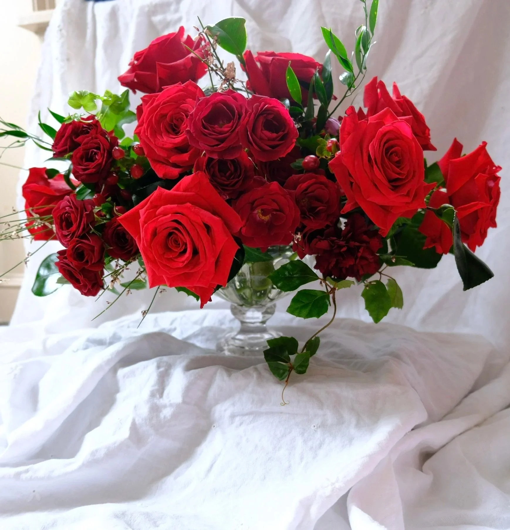 A bouquet of red roses and small red berries in a glass vase on a white cloth, with a light-colored background.