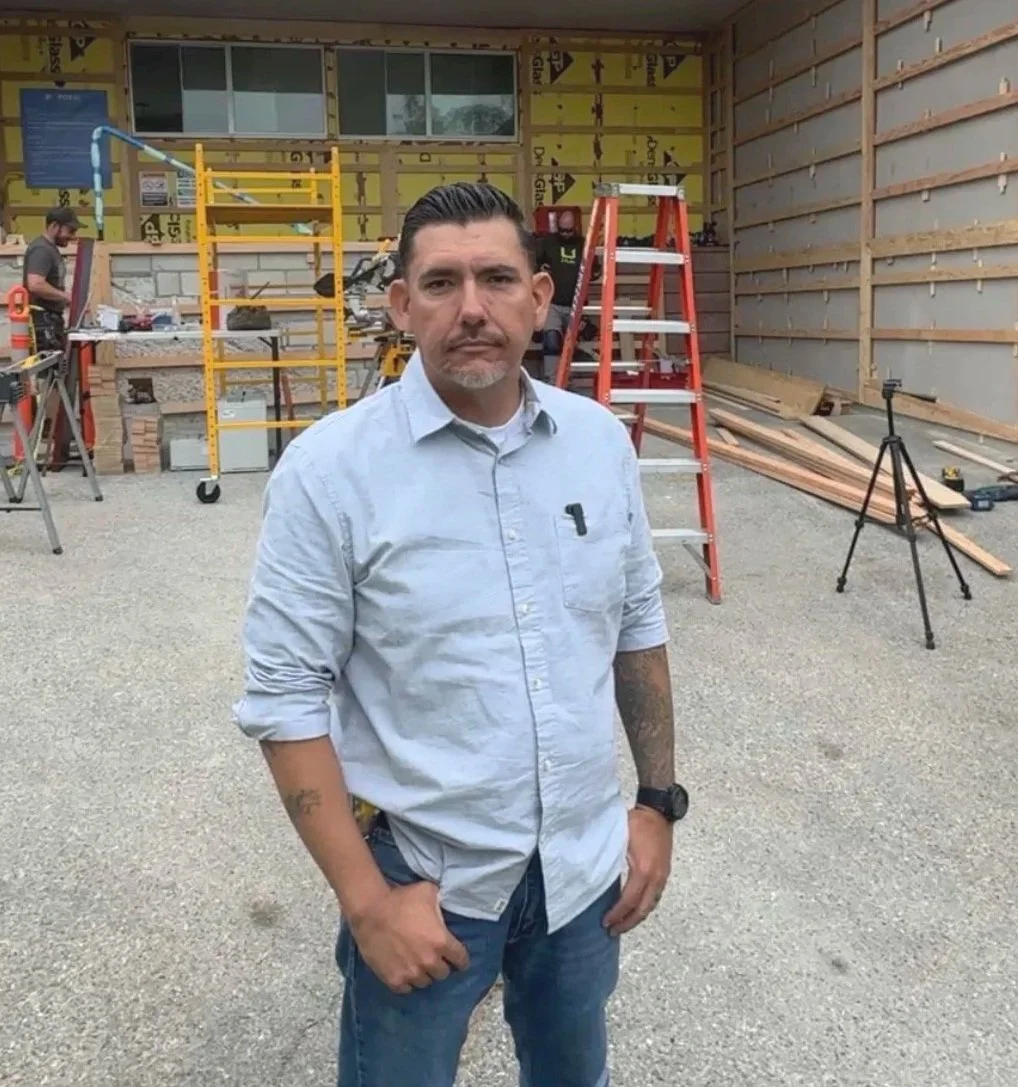 Man standing in front of a construction site with tools and ladders, wearing a light blue shirt and jeans.