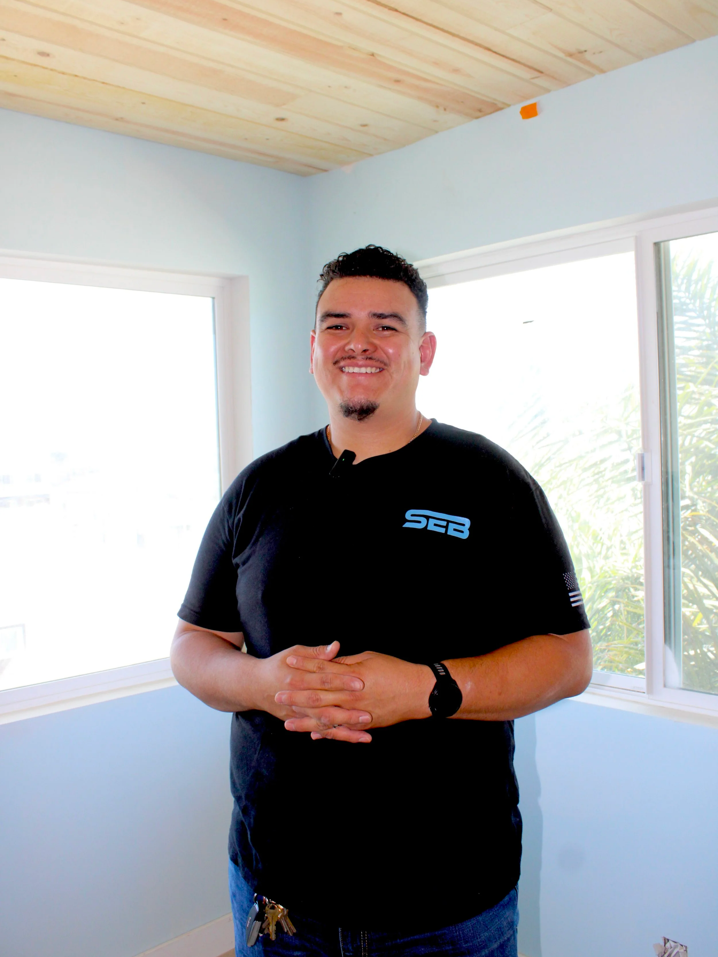 A man with dark curly hair, a goatee, and a smile, standing in a room with large windows and light blue walls. He is wearing a black t-shirt with a blue logo and has keys attached to his belt.