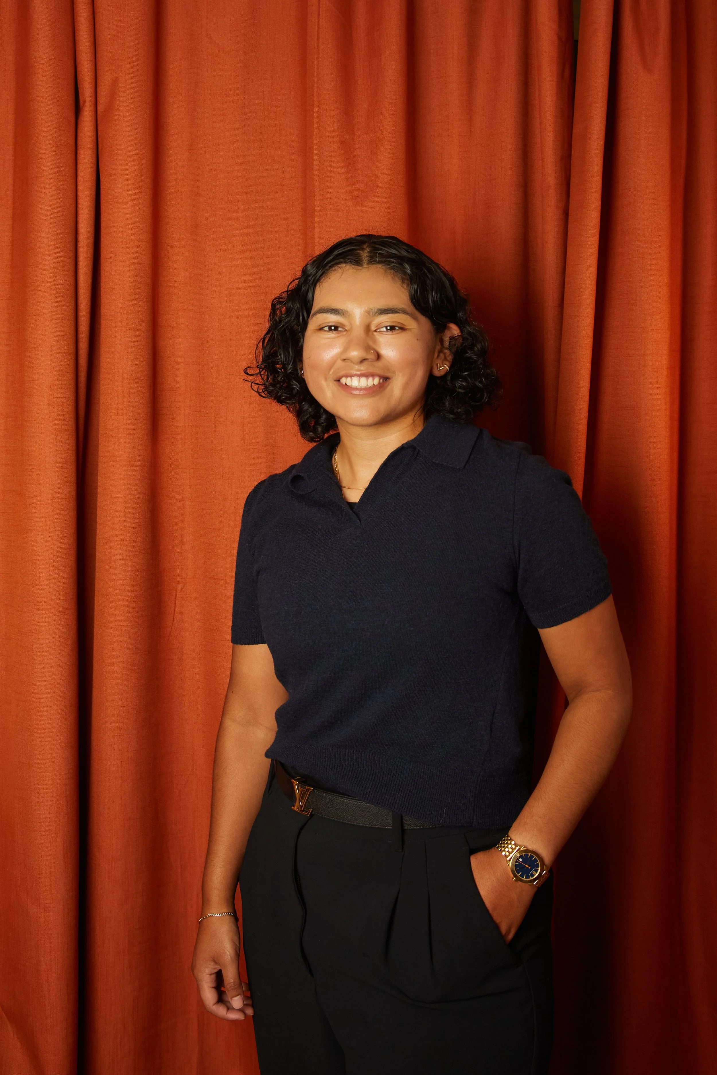 A woman with curly black hair smiling, wearing a navy polo shirt, black pants, a gold watch, and gold jewelry, standing in front of orange curtains.
