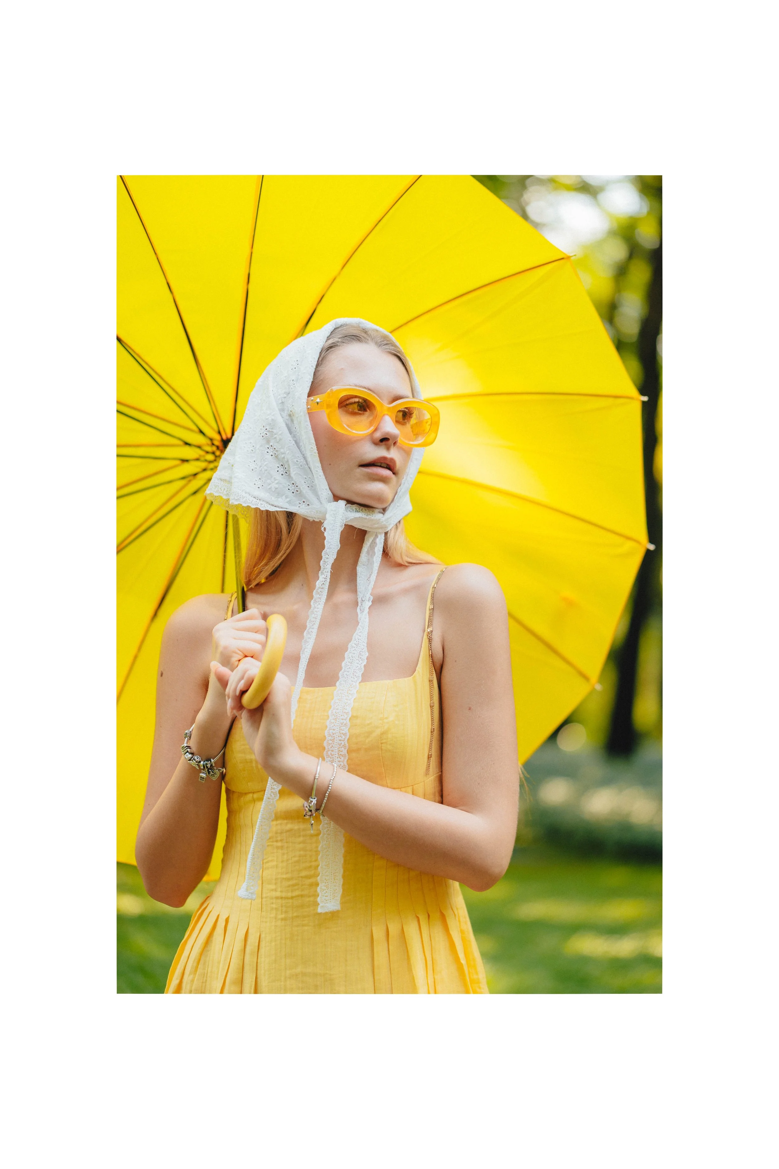 Young woman with blonde hair, wearing yellow sunglasses, a yellow dress, and a white lace headscarf, holding a yellow umbrella outdoors with greenery in the background.