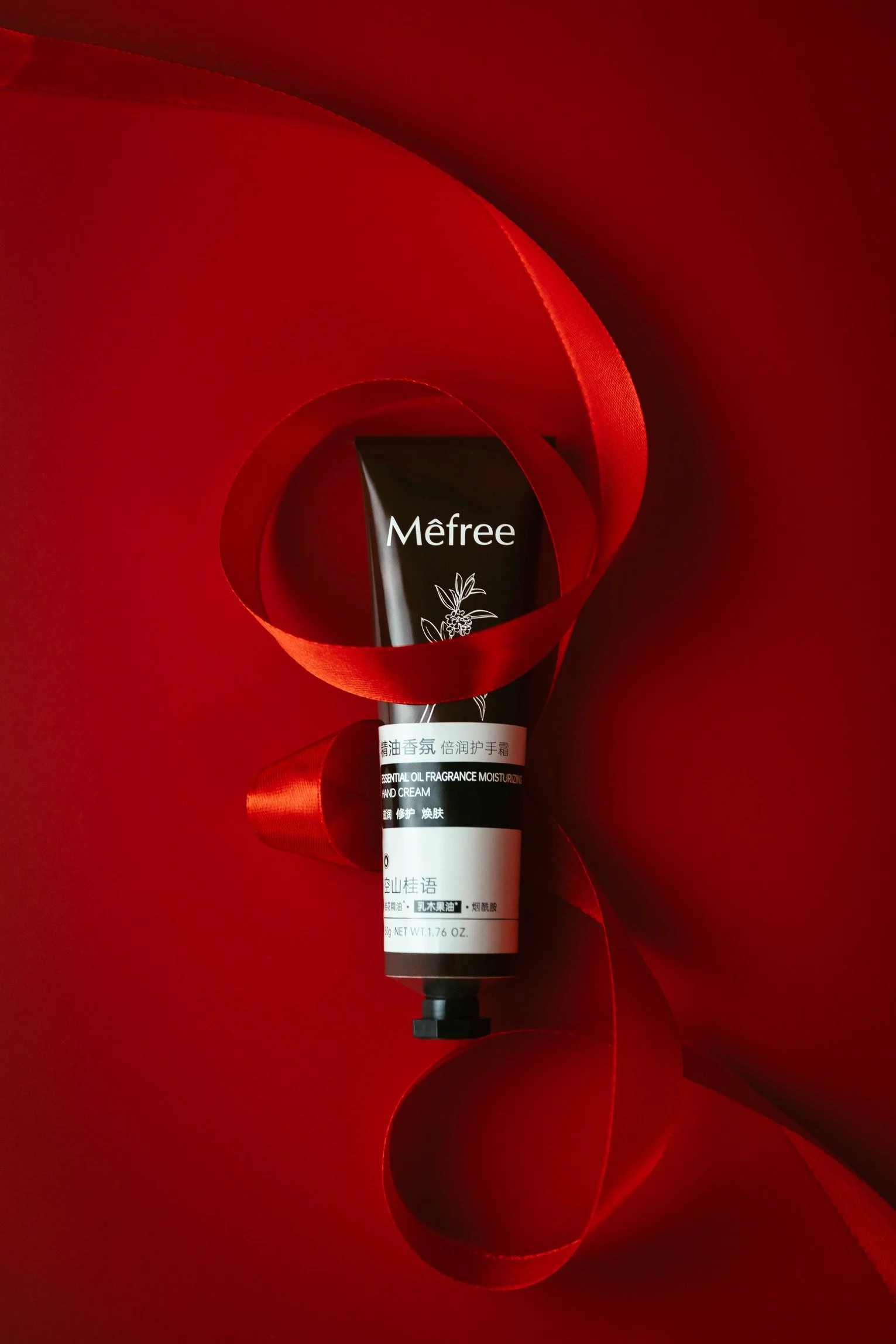 A black tube of moisturizing hand cream with floral illustrations is partially wrapped with a red ribbon, set against a red background.