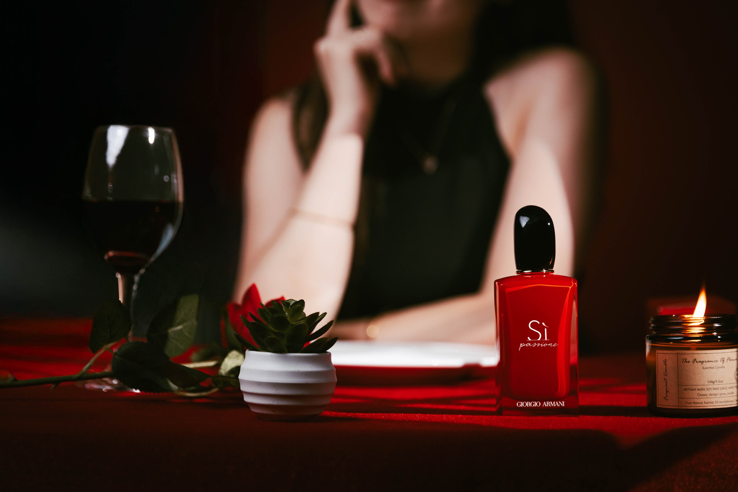 A woman in a black dress sitting at a table with a glass of red wine, a potted succulent, a red perfume bottle labeled 'Sì passione' by Giorgio Armani, and a lit candle.