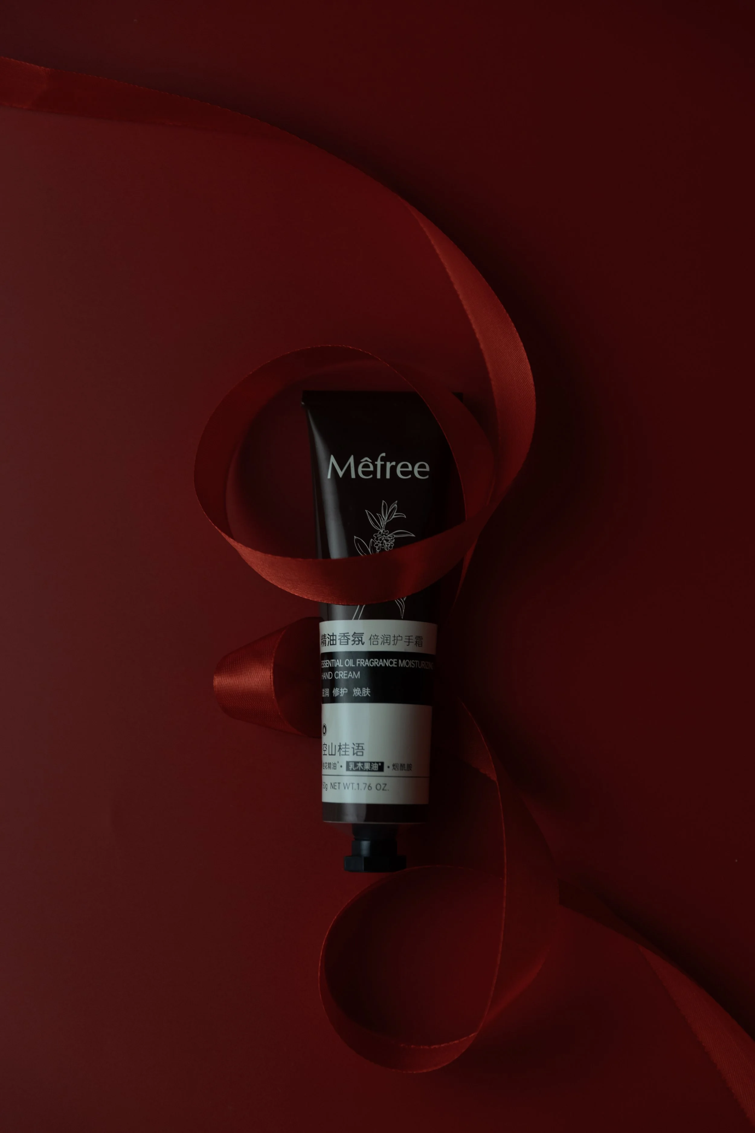 Tube of Mêfree essential oil fragrance moisturizer hand cream, partially wrapped with red satin ribbon on a dark background.