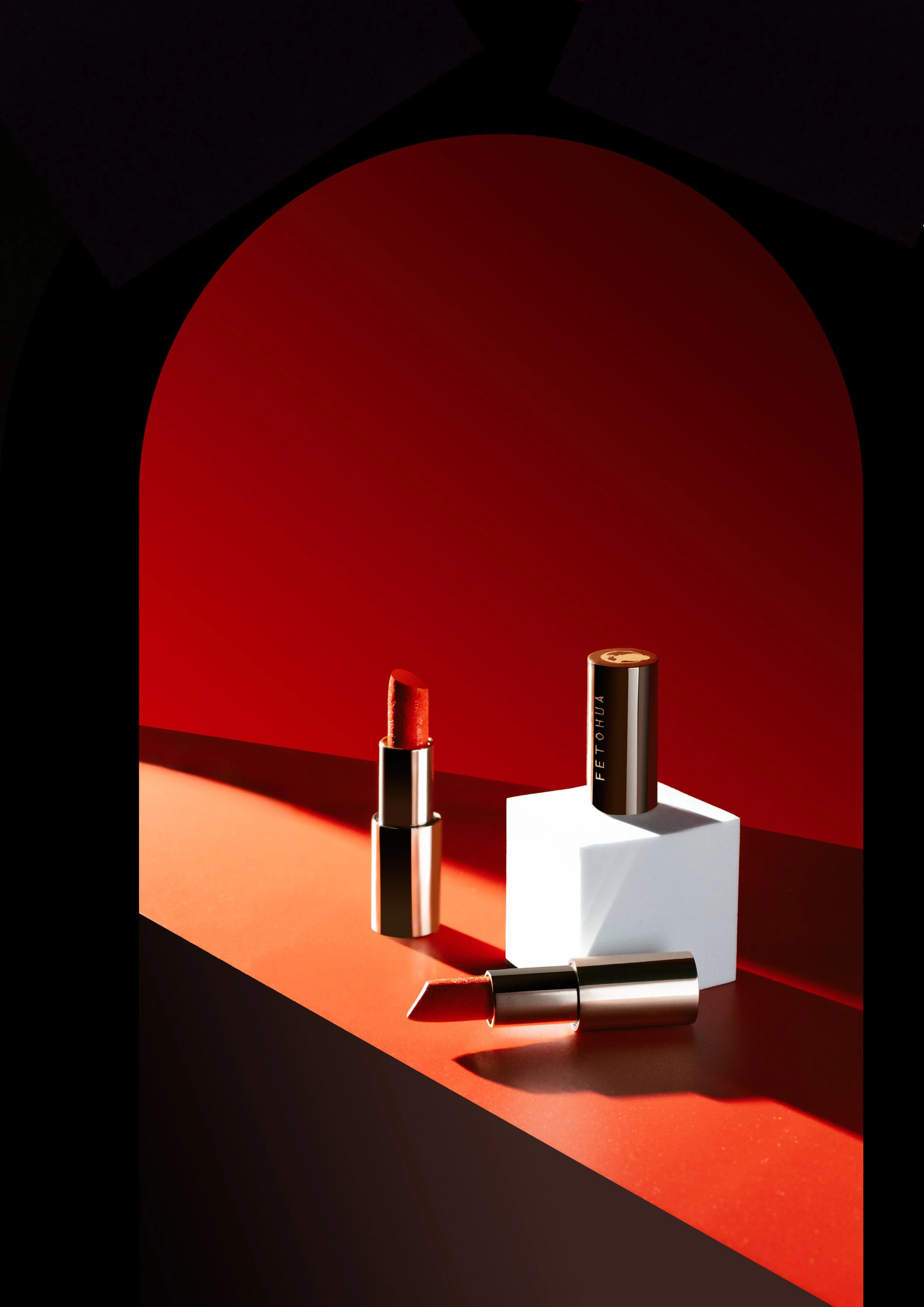 Two lipsticks—one open with red lipstick and one closed—are displayed on a red and black geometric background with a white platform, featuring dramatic shadows and lighting.