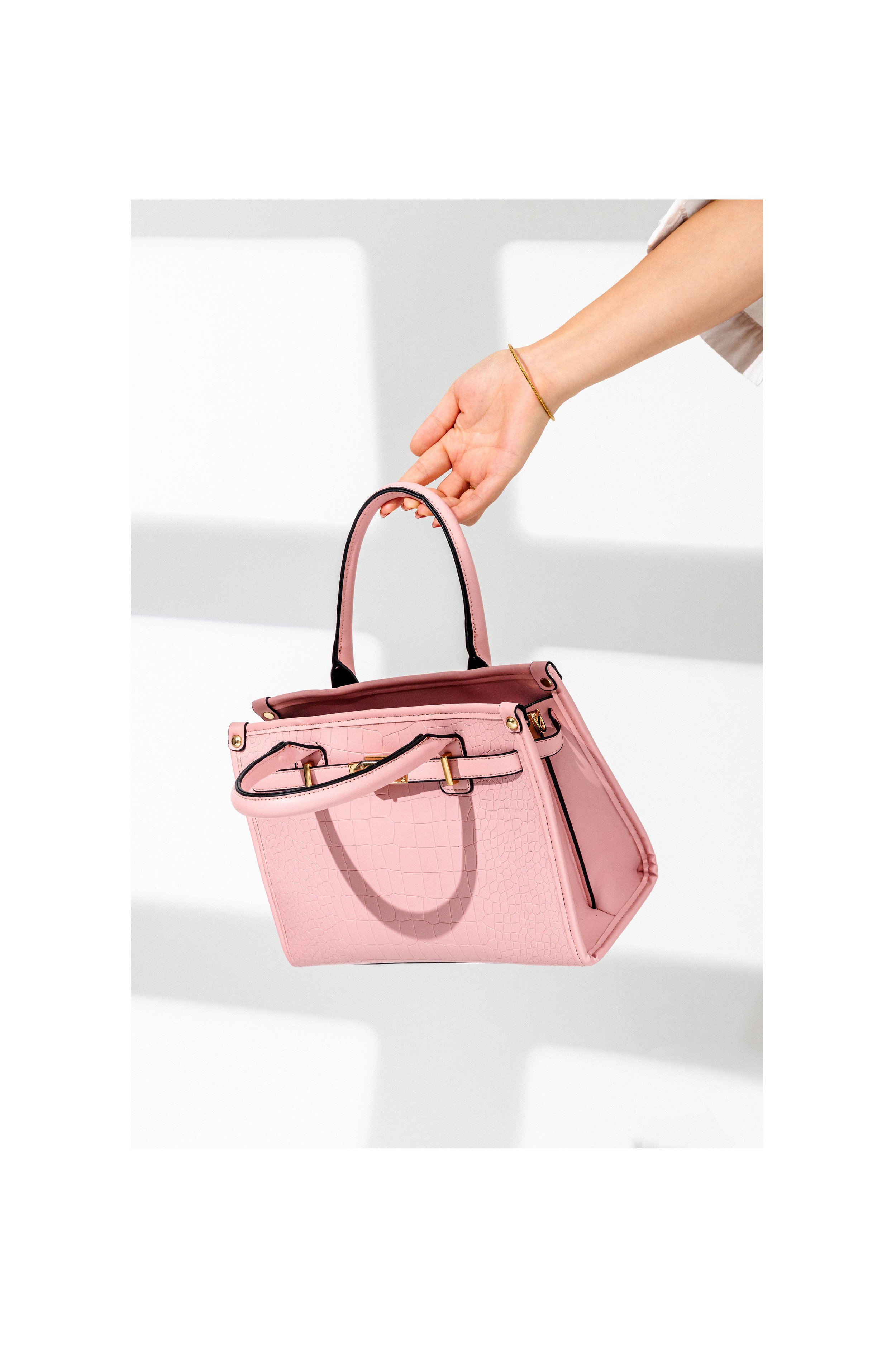 A person is holding a pink handbag with a crocodile texture, gold accents, and black trim against a white background.