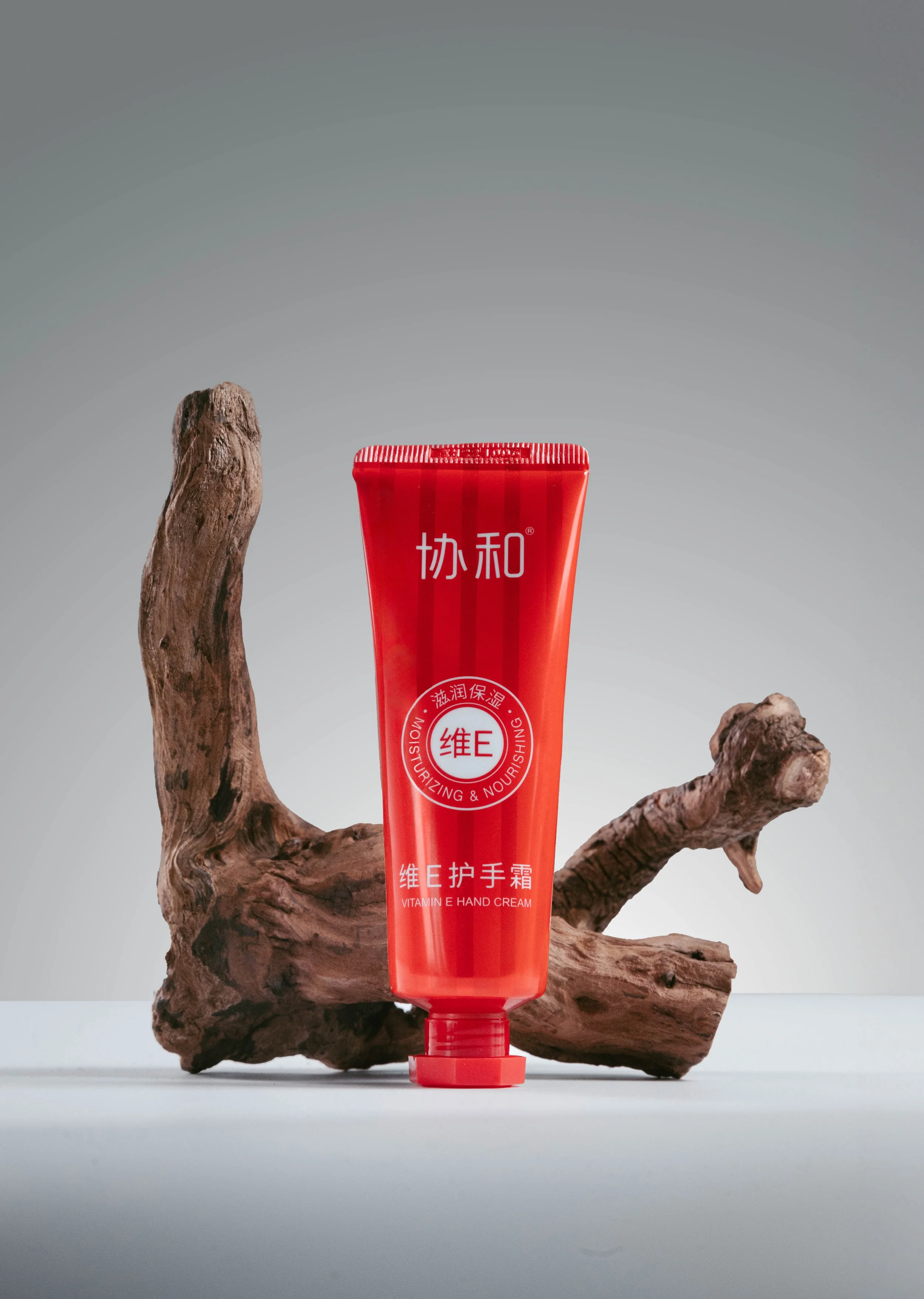 Red tube of Vitamin E hand cream placed on a white surface with a piece of driftwood in the background.
