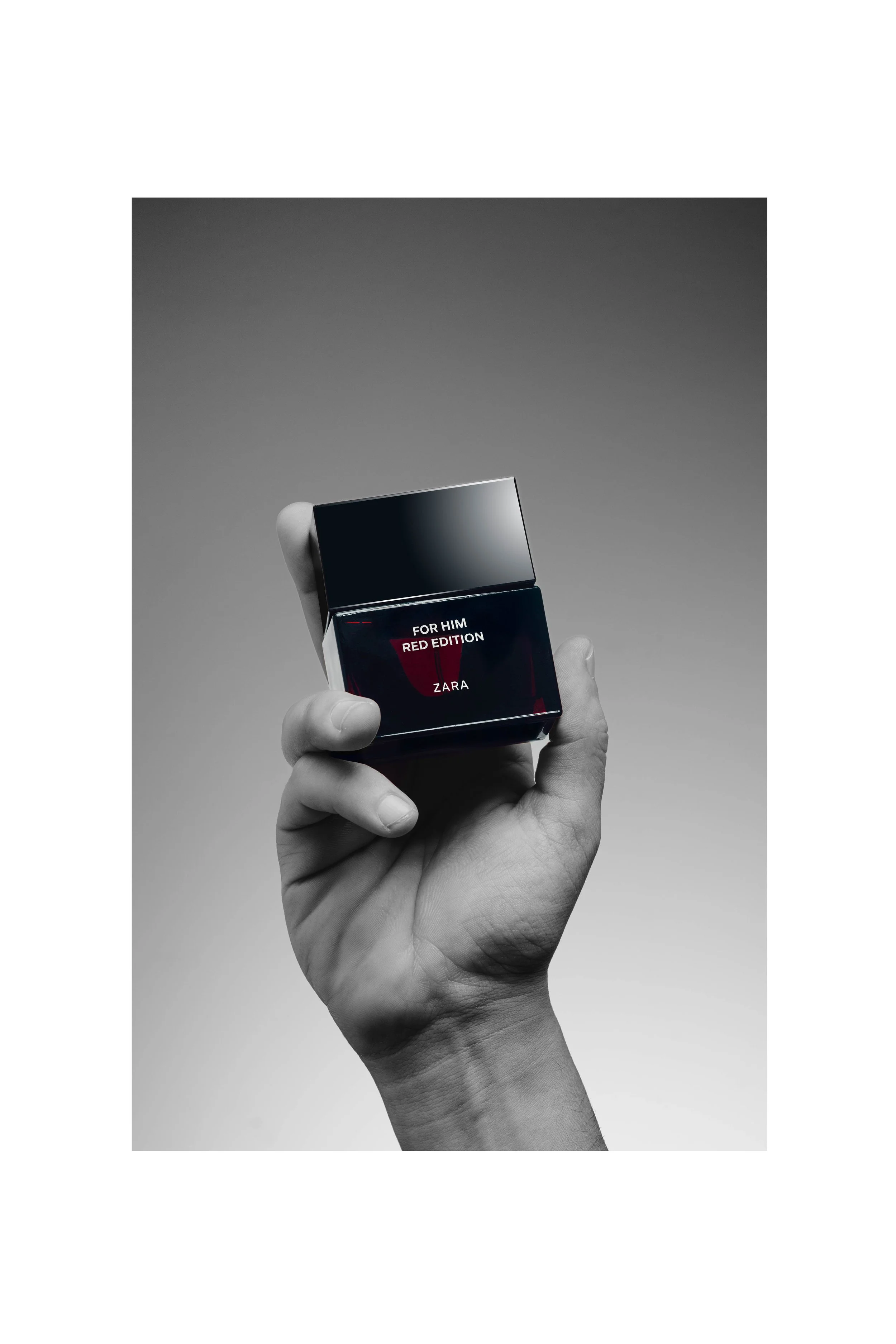 A black-and-white photo of a hand holding a small, square perfume box with a black glossy finish, with text on the box that reads "For Him Red Edition Zara."
