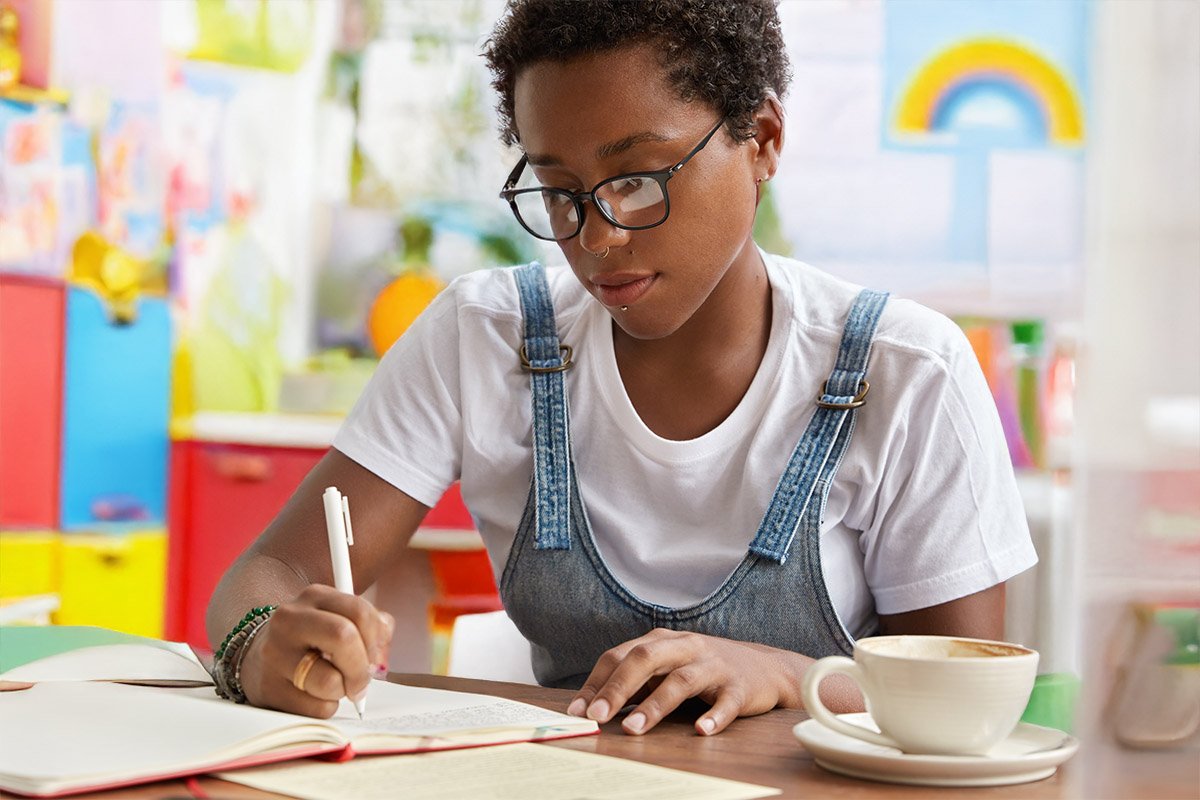 Young woman with short curly hair, glasses, and a septum piercing writing in a notebook at a colorful table in a bright room.