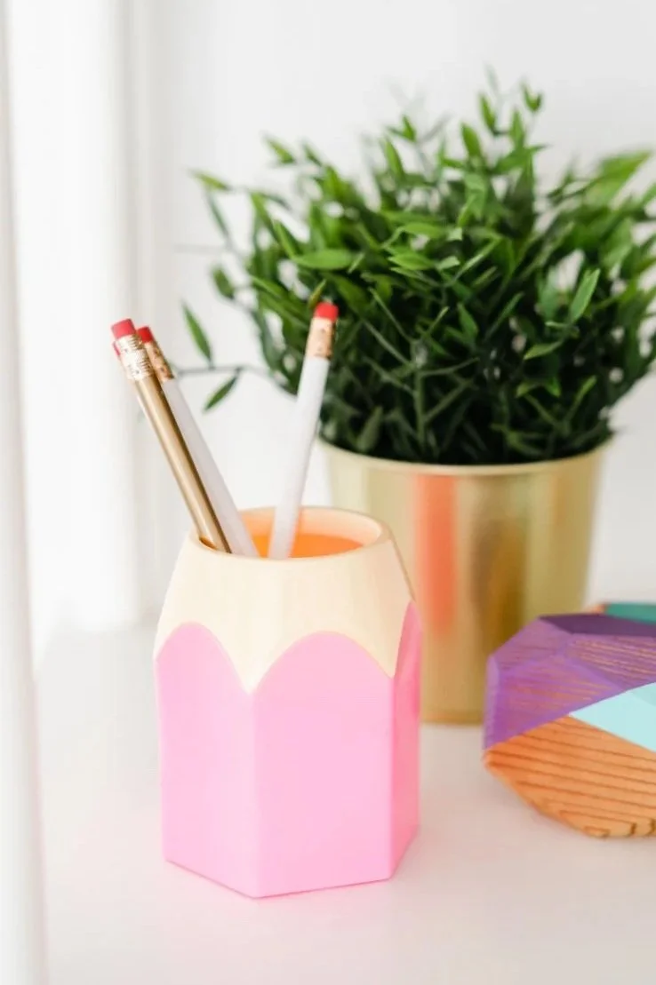 A pink and cream-colored pencil holder shaped like a peeled fruit, holding three pens and a pencil. Behind it is a potted green plant in a gold pot, and part of a colorful geometric-shaped object on a white surface.