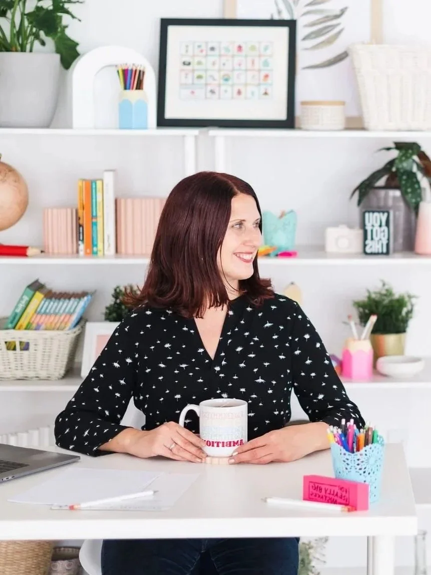 A woman with shoulder-length dark red hair is sitting at a white table, smiling and holding a mug with the word 'AMBITIOUS' printed on it. She is wearing a black blouse with white star patterns. The background features white shelves filled with colorful books, small potted plants, and decorative items, with a framed picture on the top shelf.