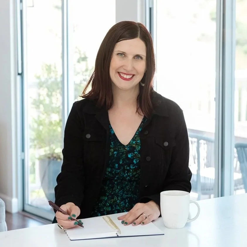 A woman with shoulder-length brown hair, smiling, sitting at a white table with a notebook, pen, and coffee mug, in front of large windows with a view of a deck and trees outside.