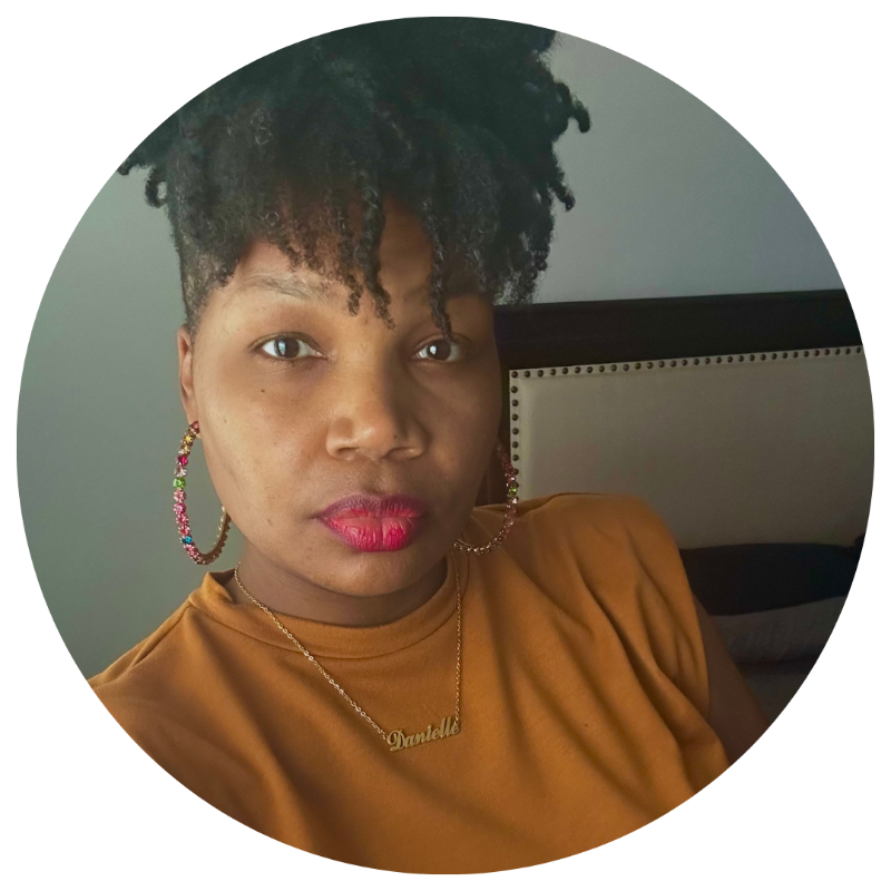 A woman with natural curly hair, wearing colorful hoop earrings, a gold necklace with the name 'Dandell,' and a mustard-colored top, sitting in front of a gray headboard.