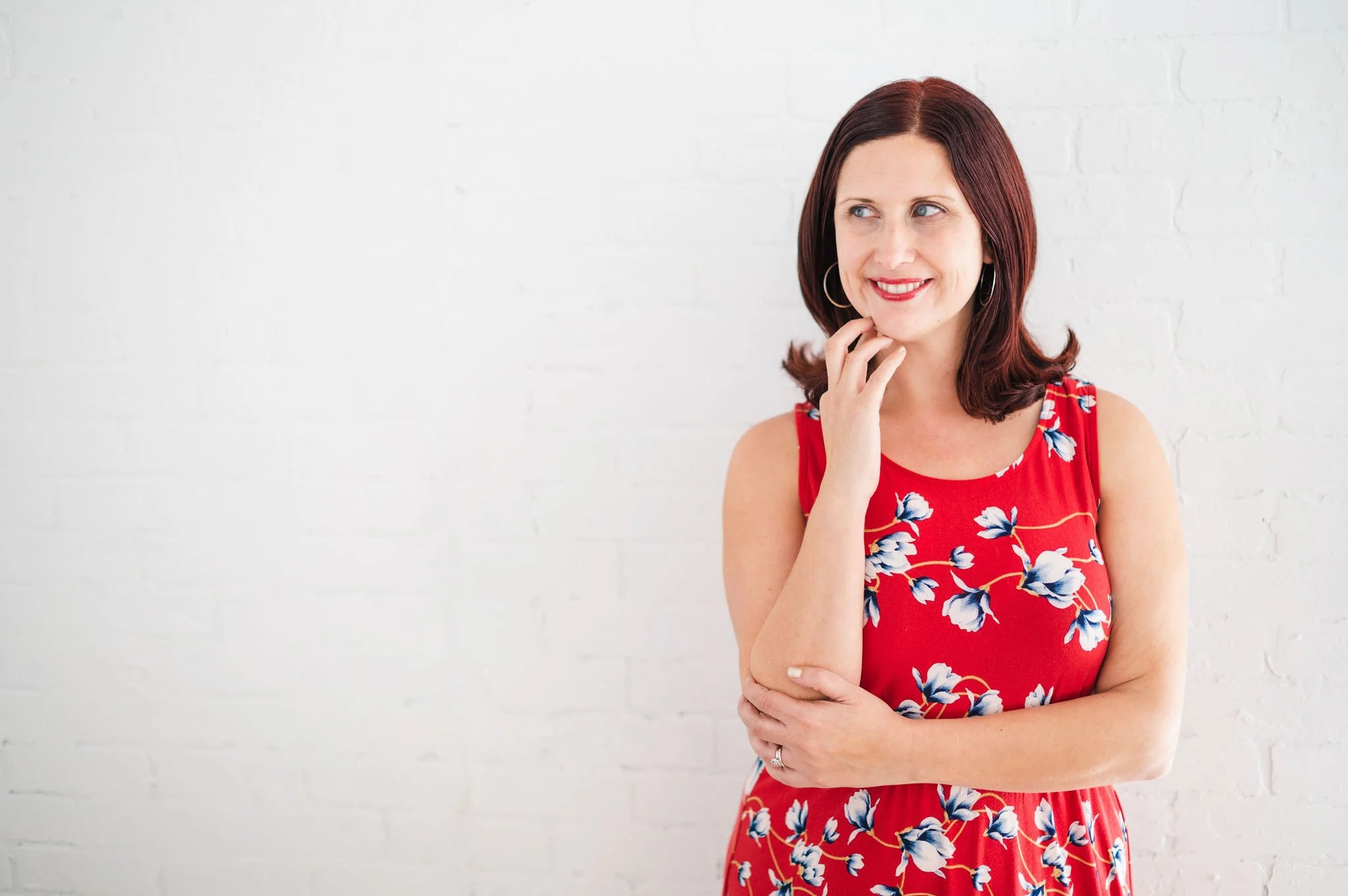 A woman with shoulder-length dark red hair wearing a sleeveless red dress with blue and white floral pattern, smiling and looking to her right, standing against a plain white wall.