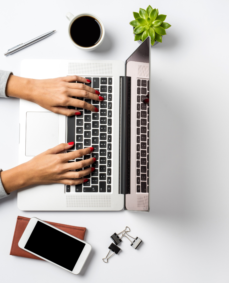 Top-down view of person typing on a silver laptop with a black keyboard, a cup of coffee, a green succulent plant, a white smartphone on a brown wallet, a pen, and paper clips on a white desk.