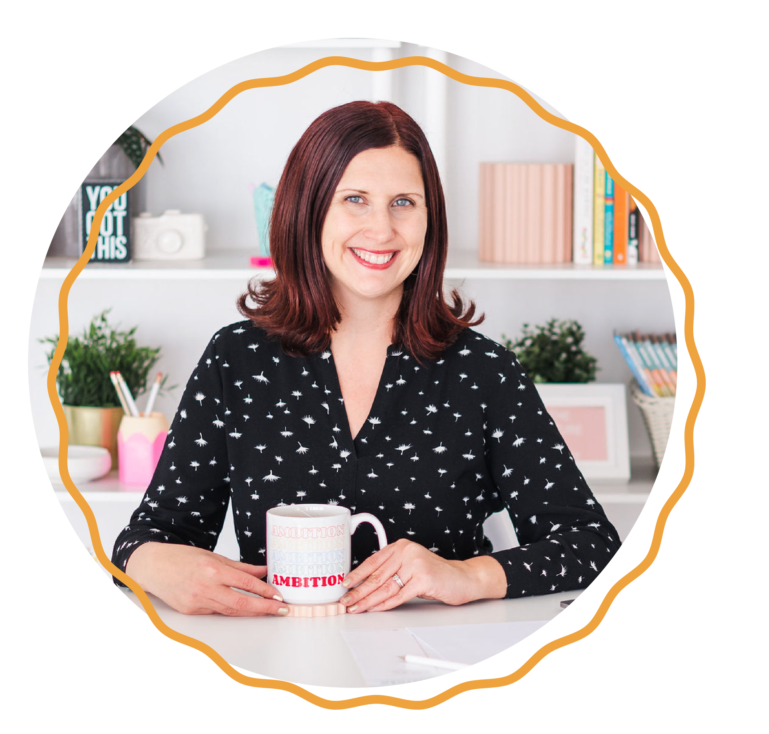Lily Jones, Educator Forever Founder, A smiling woman with shoulder-length reddish-brown hair holding a mug with the word 'AMBITION' written on it in red, sitting at a desk in a bright room with shelves and plants in the background.