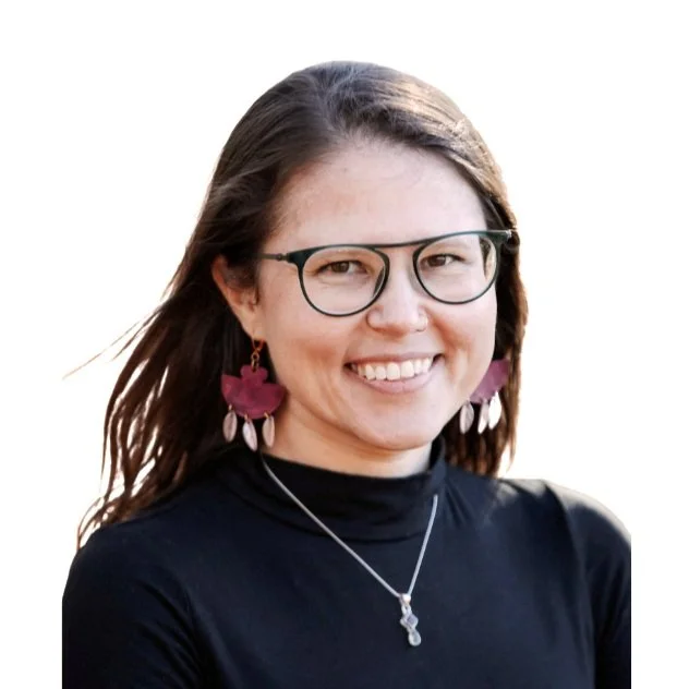 A woman with glasses, long brown hair, wearing pink and white shell earrings, a silver necklace, and a black top, smiling outdoors.
