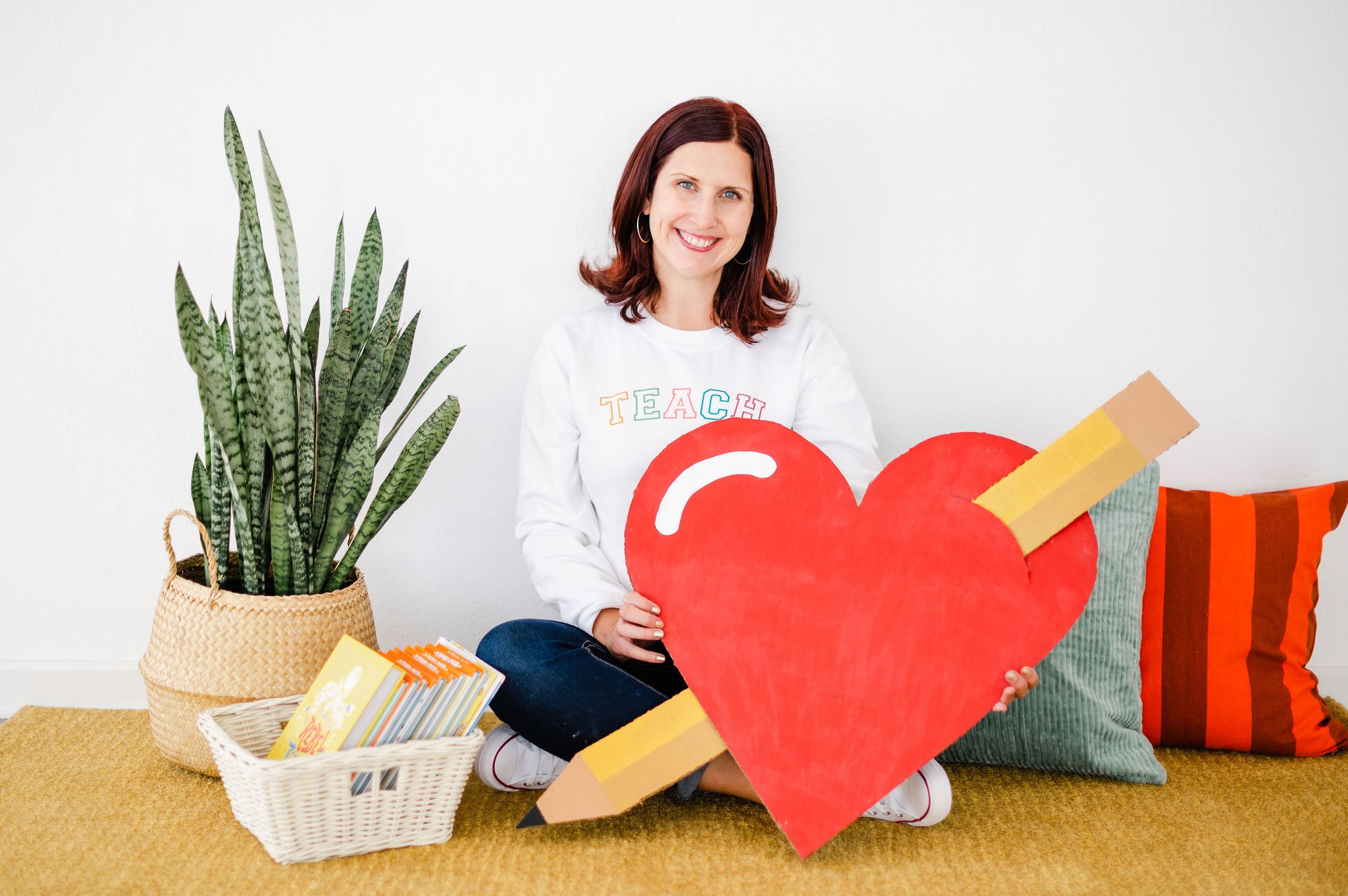 A woman sitting on a yellow carpet holding a large red heart with a yellow pencil design. She is smiling and wearing a white sweatshirt with colorful letters spelling 'TEACH'. There is a houseplant in a woven basket, a basket of books, and striped pillows in the background.