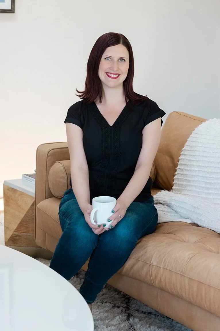 Lily Jones, Founder of Educator Forever, A woman with shoulder-length dark red hair, wearing a black top and jeans, sitting on a tan leather sofa, holding a white coffee mug, smiling in a living room with neutral decor.